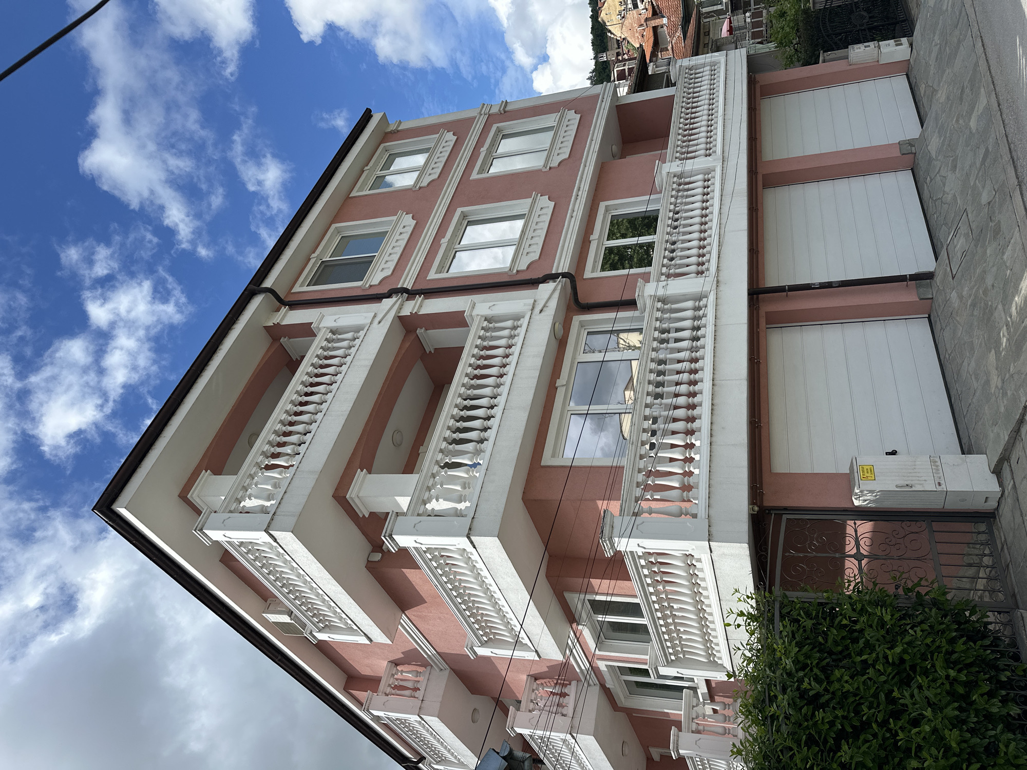 The image shows a pink and white building in Veliko Tarnovo, Bulgaria. The building has multiple stories, with windows and balconies featuring white balustrades. The balconies are positioned vertically with the balustrades on the front face of each one. The building has a black trim. The sky is blue with clouds. Part of the building includes large white doors or garage doors. Beside the building is a small gate with ornamental ironwork and a bush partially obscuring it. An electrical box is mounted to the side of the building.