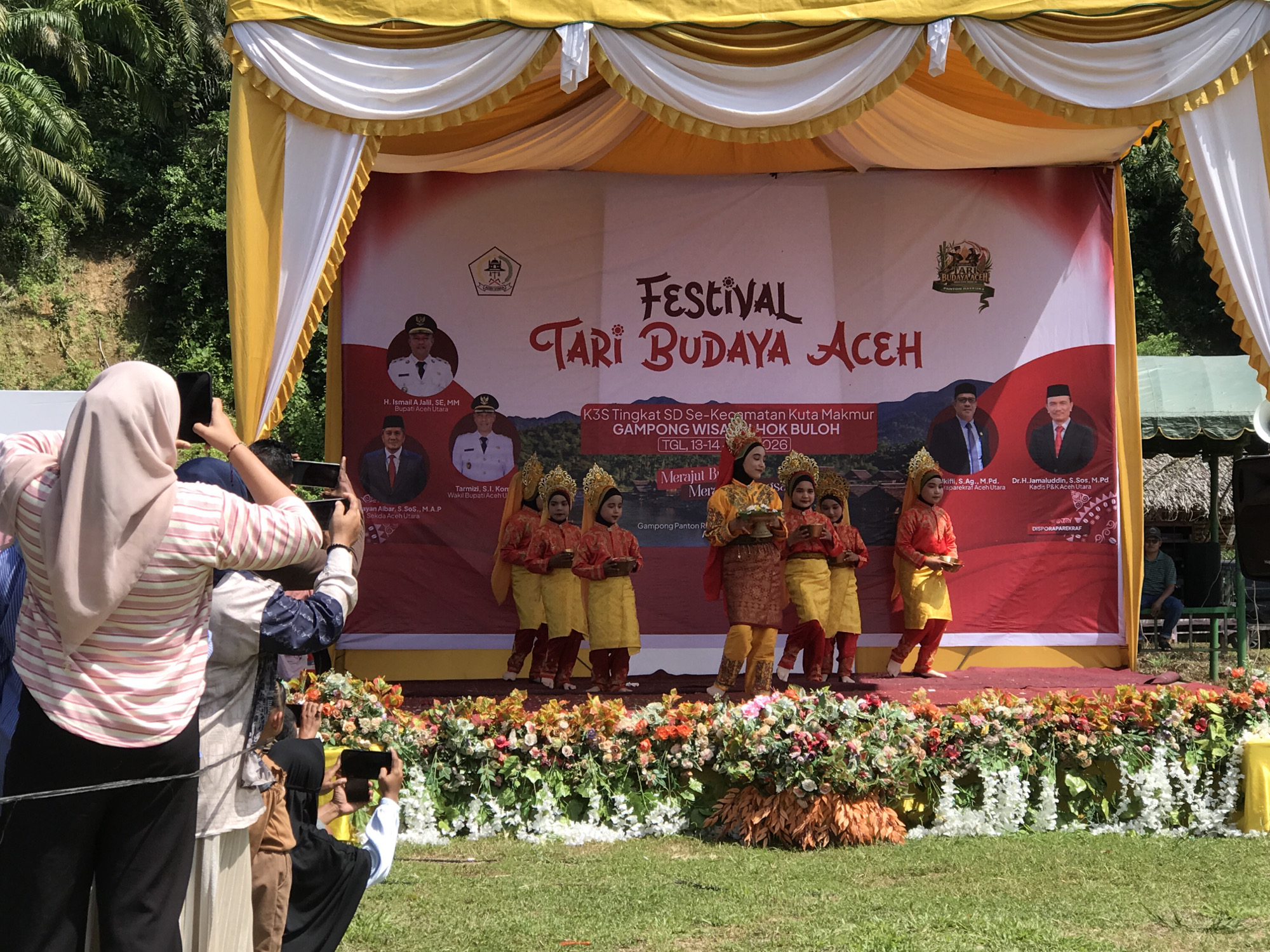 This image captures an outdoor cultural festival event. The main focus is a stage decorated with yellow and white fabric draping and adorned with a vibrant floral arrangement at its base. A large red banner serves as the backdrop, featuring event details written in Indonesian, including "FESTIVAL TARI BUDAYA ACEH" which translates to "Aceh Cultural Dance Festival." The banner also displays official seals and portraits of individuals in formal attire, likely dignitaries.
On the stage, several young performers dressed in traditional Acehnese attire are present. They wear colorful costumes consisting of embroidered tops and pants or skirts, often accompanied by decorative headwear. Some of the performers are holding small bowls.
In the foreground, a crowd of spectators is visible, some of whom are holding up smartphones to capture the event. The ground in front of the stage is covered in green grass, and lush green foliage and trees are seen in the background, suggesting a natural or semi-rural setting. The overall atmosphere appears festive and celebratory. The specific location is identified as Pantonrayeuk Satu, Indonesia, indicated by text on the banner.