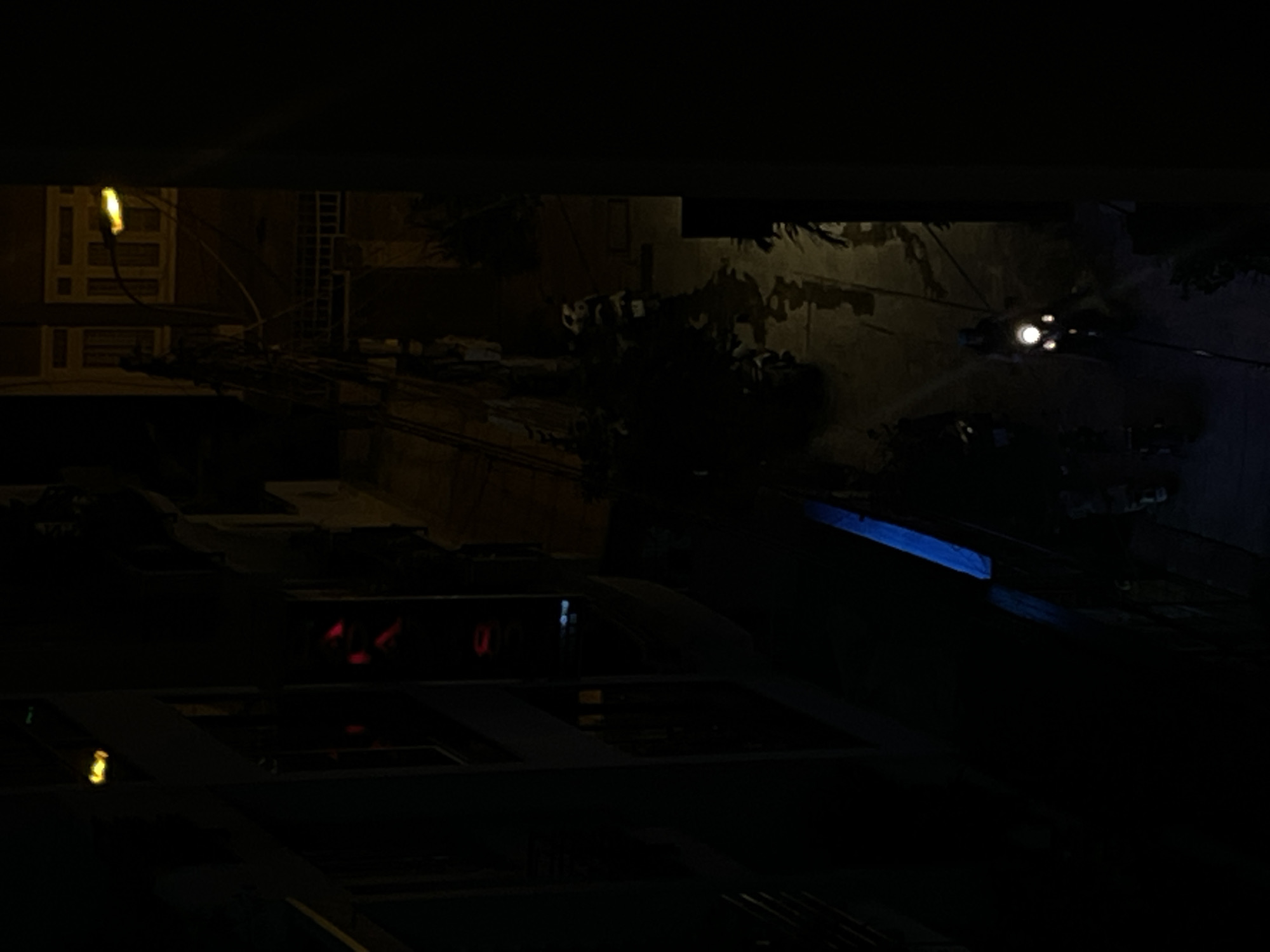 In Nha Trang Ward, Vietnam, a low-light, high-angle shot captures a dimly lit street scene at night. The foreground is dominated by darkness, with a sliver of bright blue material visible on the left. A motorcycle rider, illuminated by their headlights, is centrally positioned in the mid-ground, moving away from the viewer. The street surface appears wet, reflecting the artificial light sources. Buildings with visible windows and electric wires line the street in the background, hinting at an urban environment. Red lights are also visible in the upper left portion of the frame, possibly from another vehicle or illuminated signage.
