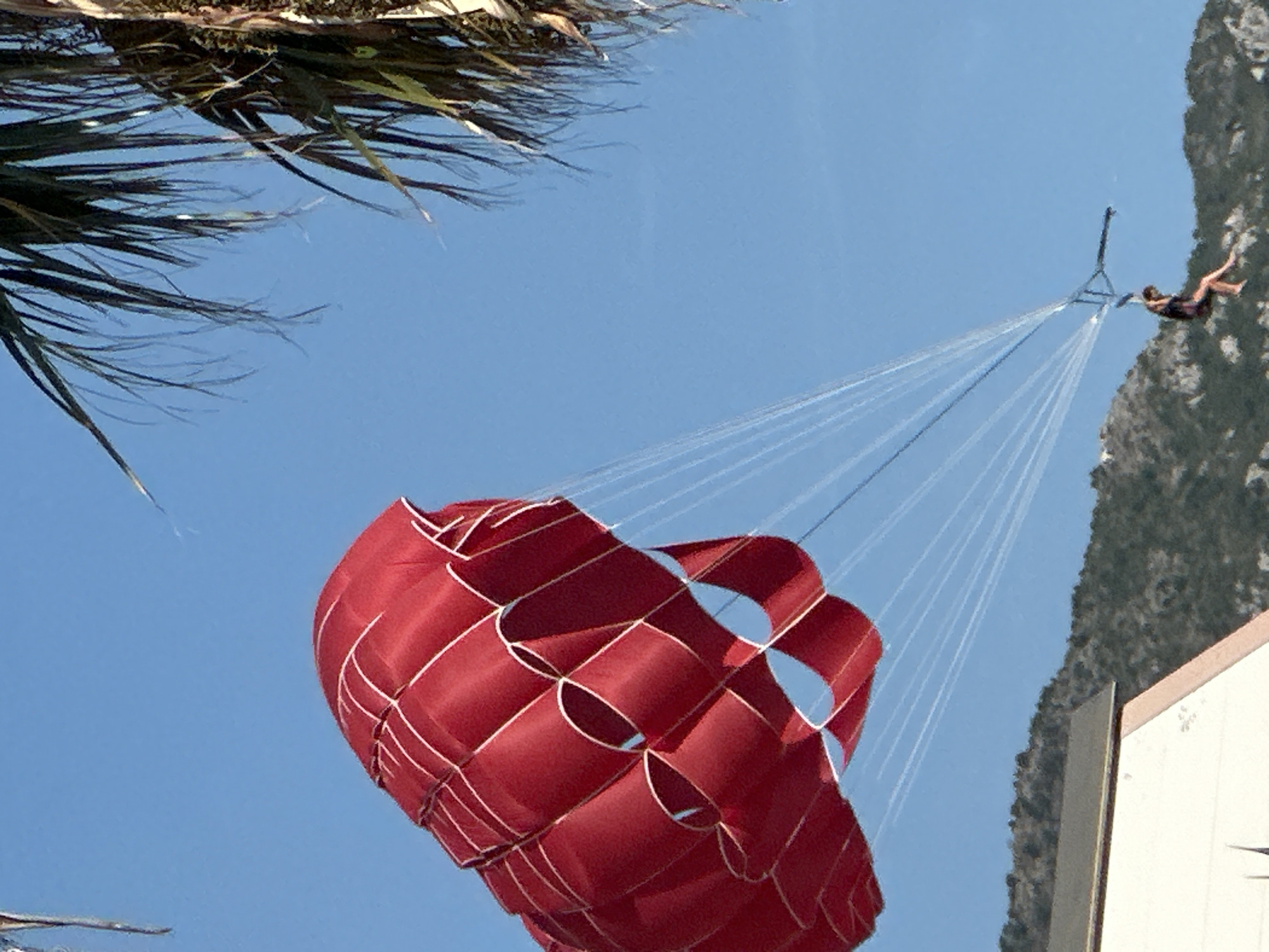 A person is parasailing on a clear blue sky day in Boreti, Montenegro. The parasail is bright red and catches the wind, creating billowing folds. The person, wearing a dark colored swimsuit, is suspended below, their legs dangling in the air, as they are pulled by a tow rope attached to a boat, which is not visible in the frame. In the foreground, a white building with a brown trim is partially visible. Behind the building, a lush green, mountainous terrain stretches across the background. The fronds of a palm tree are visible in the upper right corner of the image, suggesting a tropical or Mediterranean climate.