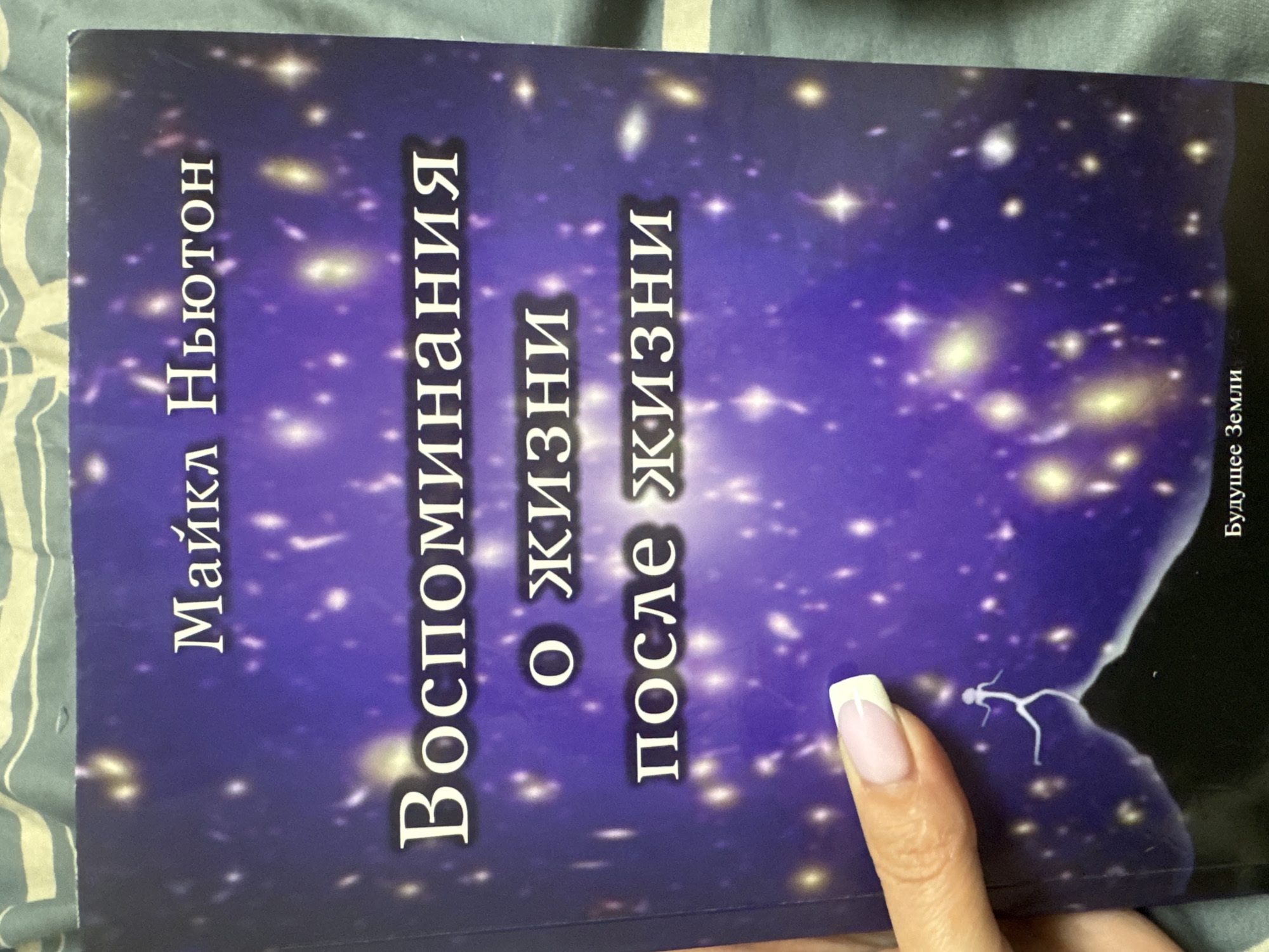 The image shows the cover of a book titled "Воспоминания о жизни после жизни" (Memories of Life After Life) by Майкл Ньютон (Michael Newton). The cover features a starry, cosmic background in shades of deep purple and blue, with glowing stars and nebulae. In the foreground, a woman's hand with a French manicure is visible on the left, touching the book's surface. At the bottom of the cover, there is a silhouette of a small, stick-figure-like person running. Below the silhouette, text reads "Будущее Земли" (Future of Earth). The book is placed on a striped fabric, possibly a bedspread or blanket, in green and white. There is no discernible location in the city of Komsomolsk-on-Amur, Russia, indicated in the image.