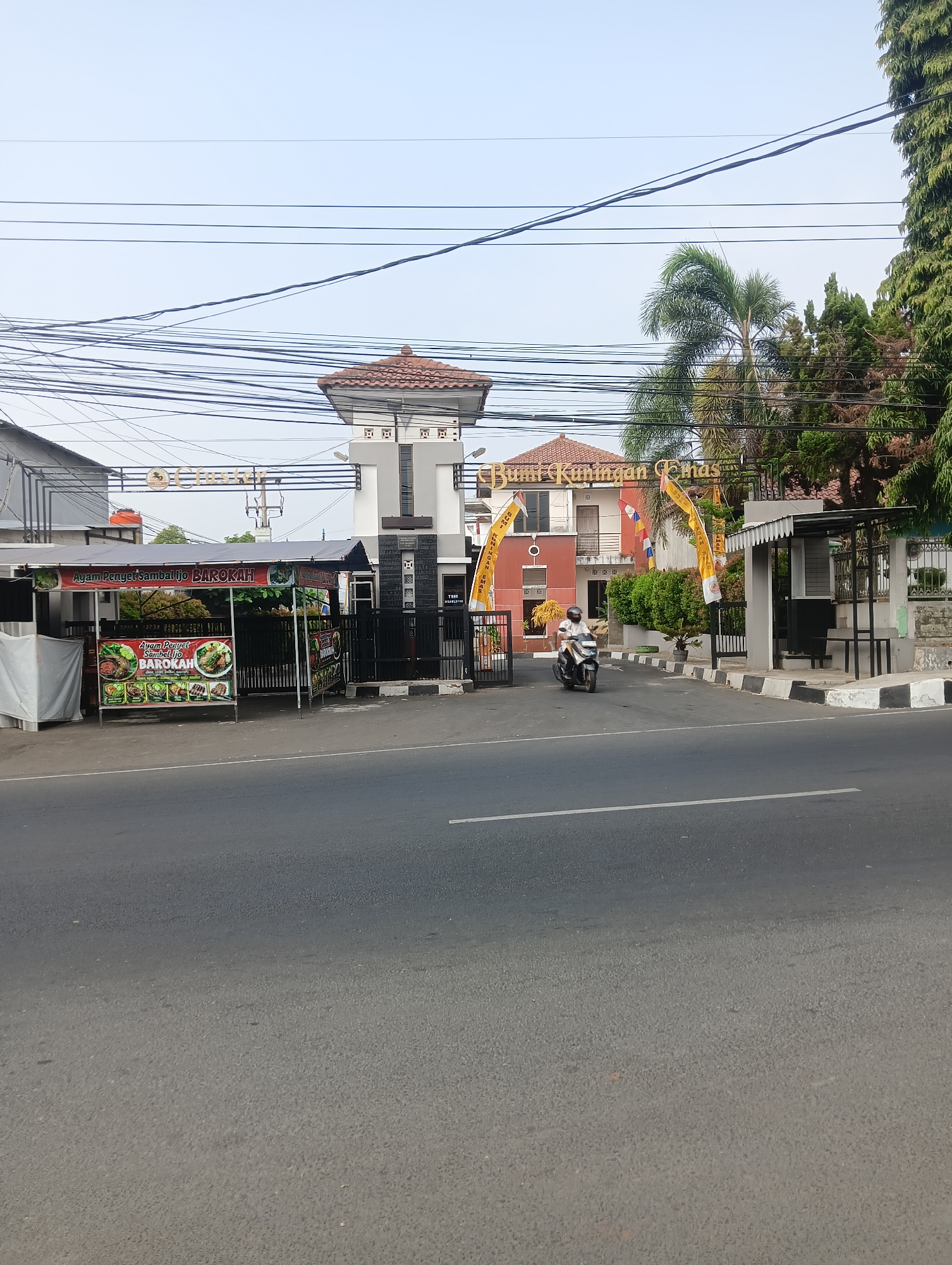The image shows a street scene in Ciporang, Indonesia. The foreground features a paved road with a white line marking a lane. On the left side of the road, there is a small food stall with a banner displaying pictures of food and the text "Ayam Penyet Sambal Ijo BAROKAH". Behind the food stall, there is a white structure, possibly a wall or building.

Further down the street, there is an entrance to a complex. A tall, white gate structure with a red tiled roof stands at the entrance, flanked by black gates. Beyond the gate, a building with red and orange walls and a red tiled roof is visible. The name "Bumi Kuningan Emas" is displayed prominently on the building in golden letters. Yellow banners with Indonesian text hang on either side of the entrance.

A person on a motorbike is entering the complex from the street. To the right of the entrance, there is a small, covered waiting area with a bench. Trees and a tall conifer tree are visible in the background, along with a clear sky with numerous power lines crisscrossing it. The overall scene depicts a typical street view in an Indonesian town, with commercial establishments and residential or institutional buildings.
