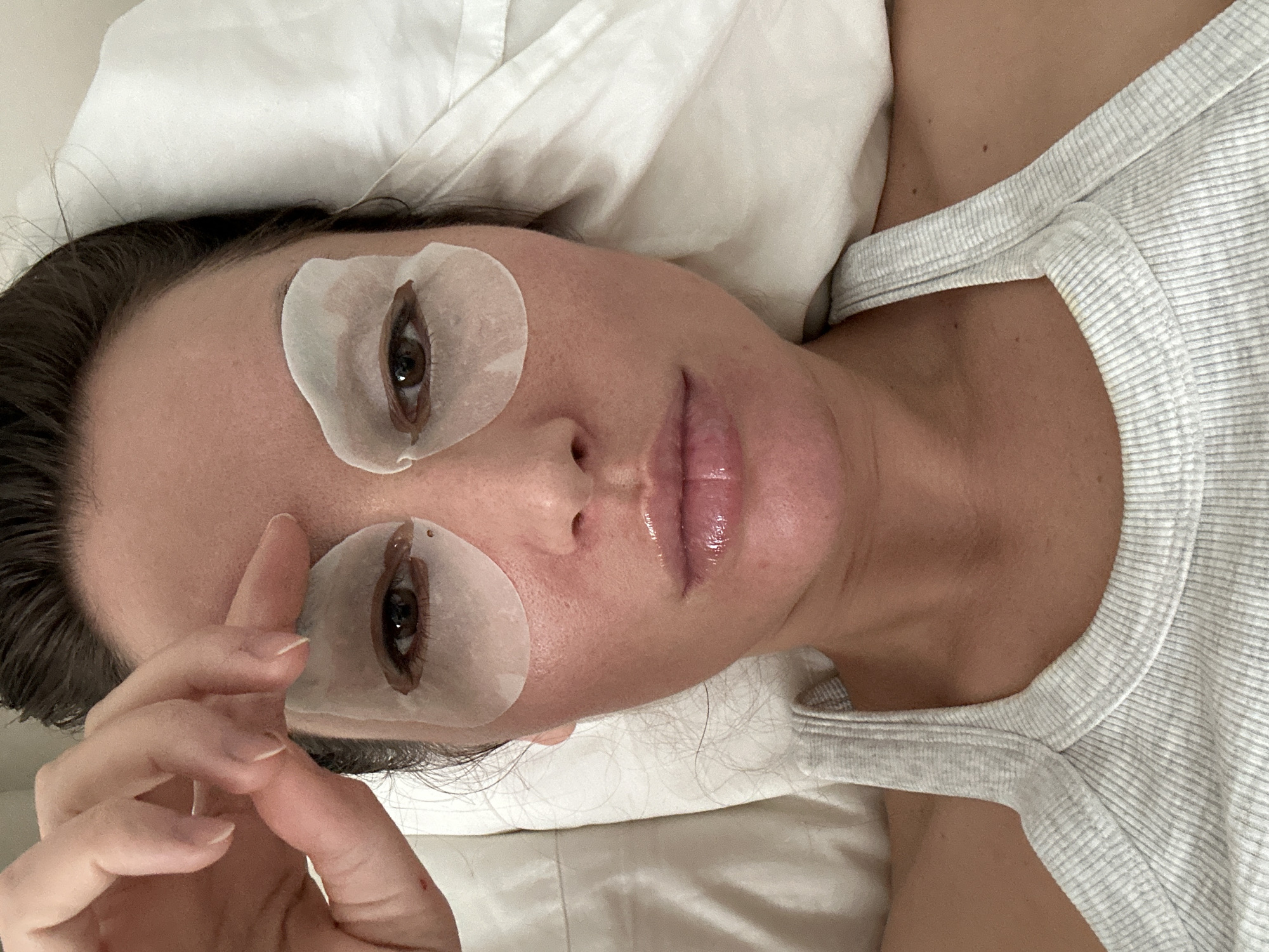 The image displays a person lying down, viewed from a slightly elevated, tilted perspective. The subject, a person with dark hair, looks directly at the camera with a neutral expression. White, translucent eye patches are positioned under each of their open brown eyes, with dark eyelashes visible beneath the patches. Their lips appear full and glossy, possibly from lip gloss or balm.
The person is wearing a light gray, horizontally ribbed tank top or camisole. Their head rests on a soft, white pillow, suggesting an indoor setting like a bed or a treatment table. One hand is partially visible, with fingers gently curled and resting near their temple.
The presence of the eye patches indicates a self-care or beauty routine in progress, such as an eye mask application or preparation for an eyelash service like extensions or a lift. The overall atmosphere conveyed is one of calm and relaxation, implying a moment of personal pampering or rest. No discernible text is visible within the image. This scene takes place in Moscow, Russia.