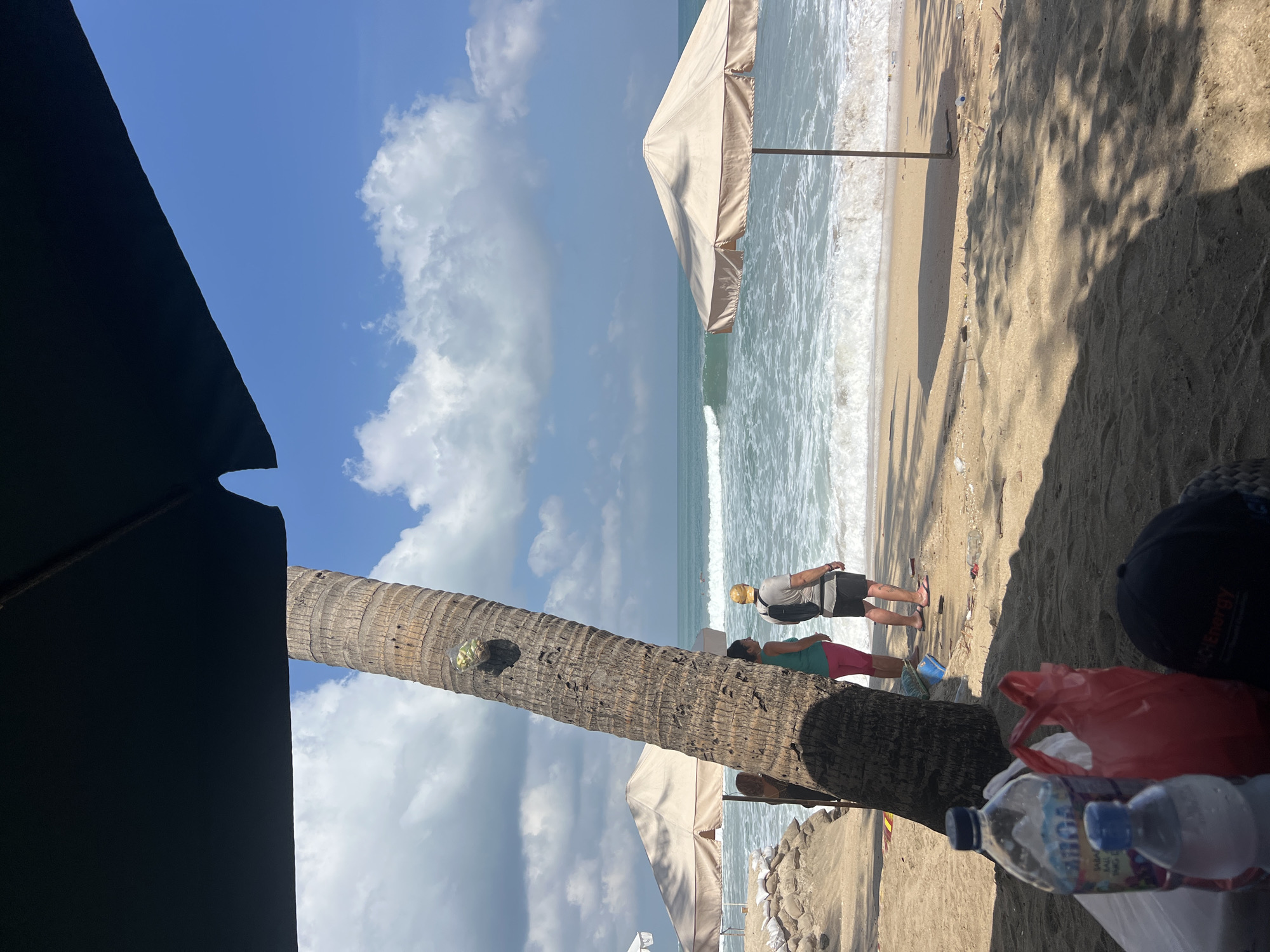 This image captures a beach scene in Kuta, Indonesia. In the foreground, two water bottles and a red plastic bag are placed on the sand. A dark baseball cap rests in the lower right corner. A large palm tree dominates the left side of the frame, with a clear plastic bag containing what appears to be fruit hanging from it. Two people stand near the palm tree, facing the ocean. One person, wearing a green tank top and pink pants, stands to the left, while the other, wearing a grey t-shirt, dark shorts, and a yellow cap, stands to the right. Both are facing the water.
In the background, the ocean features moderate waves rolling onto the shore. Two beach umbrellas with light-colored canopies are visible, one closer to the palm tree and the other further to the right. The sand is a light brown, and the sky is a bright blue with scattered white clouds. The overall impression is a relaxed beach day with visitors enjoying the coastal scenery.