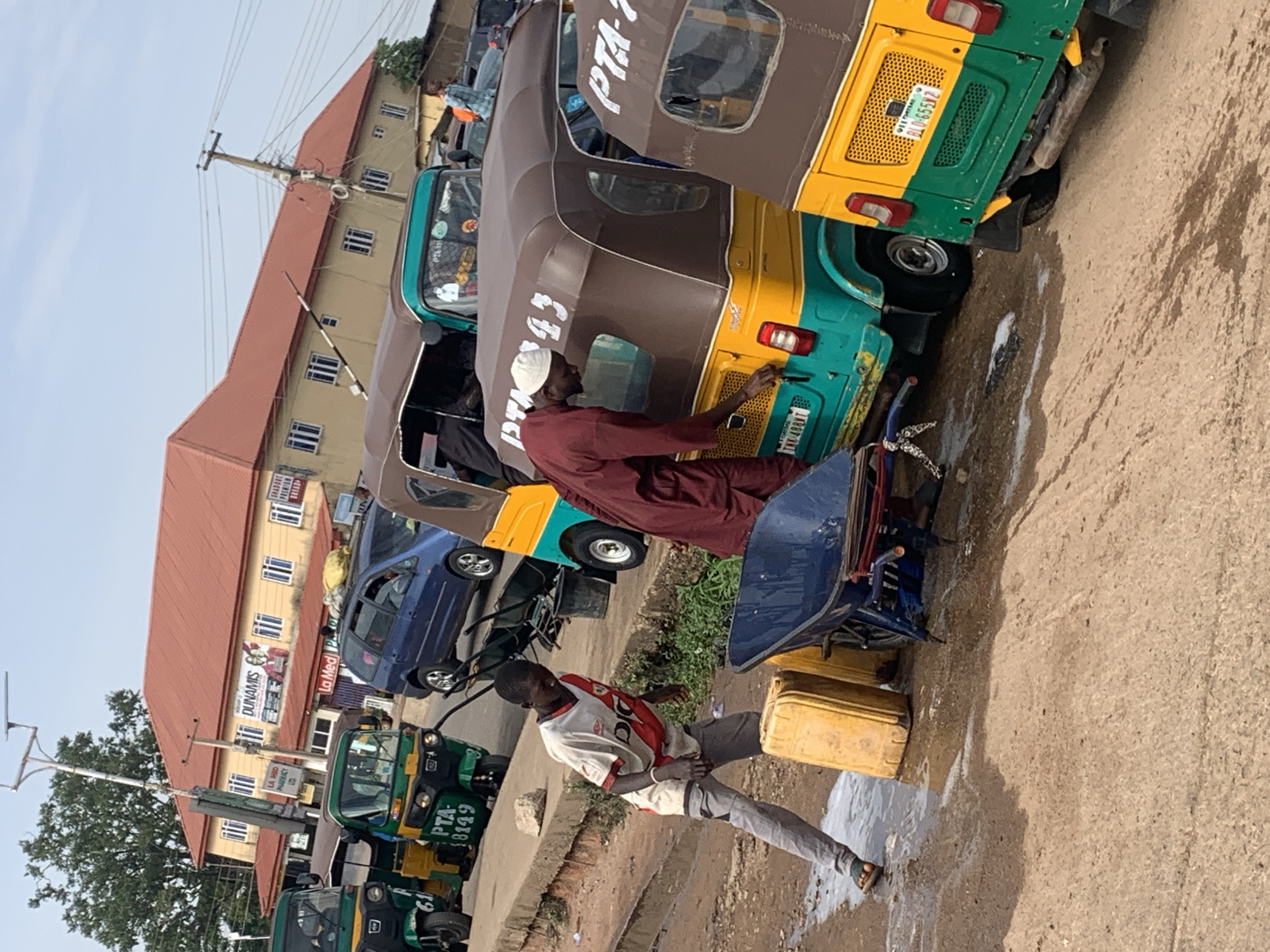 In Gura Top, Nigeria, an outdoor scene depicts a bustling street with several auto-rickshaws, also known as Keke Napep. One young man, wearing a white and red t-shirt and gray pants, is actively engaged with two large yellow water containers. Beside him, a blue wheelbarrow is positioned, hinting at some sort of task or transport. In the background, two Keke Napep vehicles, one predominantly brown and teal with "PTA 43" visible on its side, and another mostly yellow and teal with "PTA-2" on its side, are parked. A man in a maroon robe and white cap is seen interacting with the brown and teal Keke Napep, possibly closing its trunk. Further in the background, a blue minivan is parked, and a building with a red roof and signage for "DUNAMIS PHARMACY" stands prominently. The ground is a mix of dirt and puddles, suggesting recent water activity. The sky is clear and the overall atmosphere appears to be that of a busy commercial or transport hub.