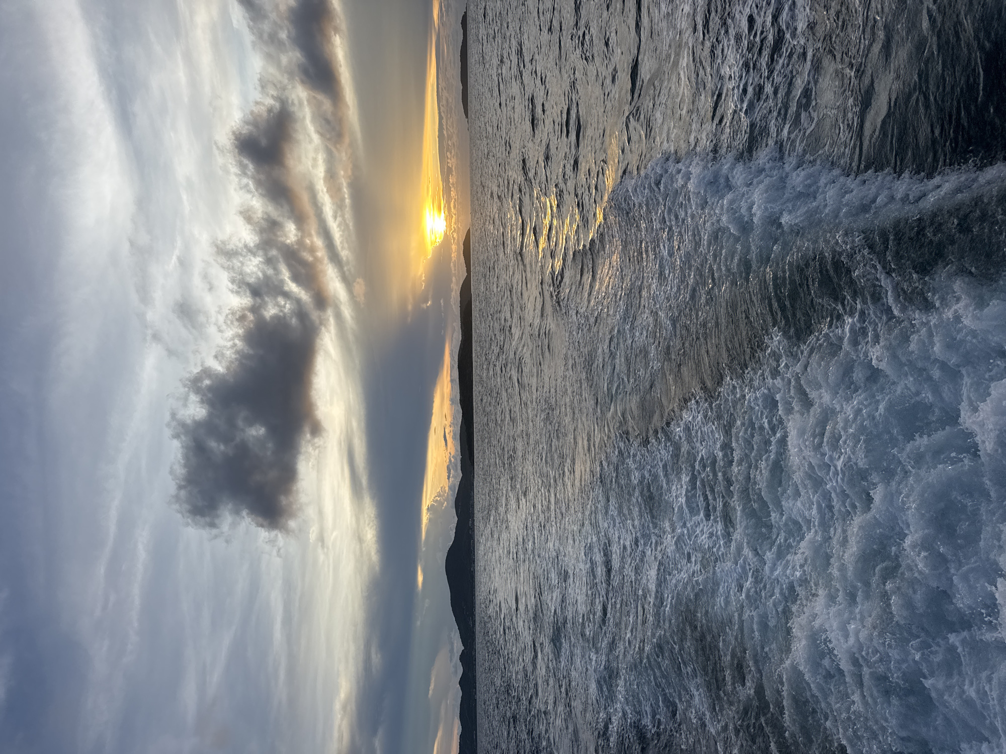 The image shows a sunset over the ocean. The sky contains clouds in various shades of gray and white. The sun is positioned low on the horizon, with a mountain range visible in the distance. The ocean surface is reflecting the sunlight, and there are waves in the foreground. It appears to be taken near Pattaya City, Thailand based on the presence of sea and mountains. There are no people visible in the image. The main subjects are the sky, the ocean, and the sunset. The photograph captures the natural environment at sunset.