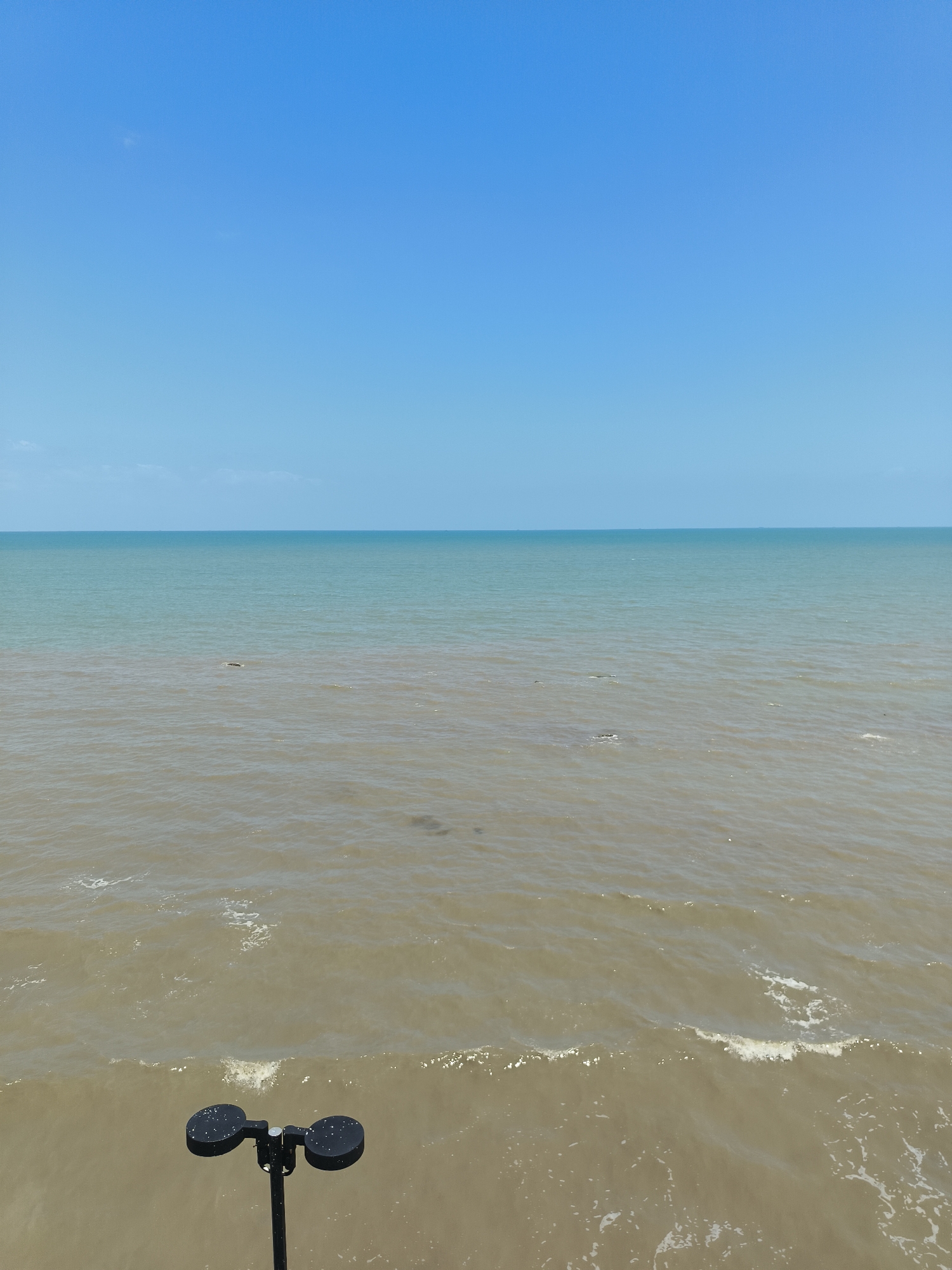 The image captures a serene coastal scene under a clear blue sky. The ocean occupies the majority of the frame, divided into two distinct color zones. The upper portion of the water is a clear turquoise, suggesting deeper, cleaner water, while the lower portion is a murkier, sandy brown, indicating shallowness or sediment. The two water colors meet in a visible line across the horizon. Small waves are present, creating a gentle ripple effect on the surface of the water. In the foreground, a black pole with two circular, black anemometer cups is visible, angled slightly to the right. The background consists of the vast expanse of the blue sky, with a hint of a light cloud formation on the horizon. The overall impression is one of natural tranquility. The location is identified as Beringin, Indonesia.