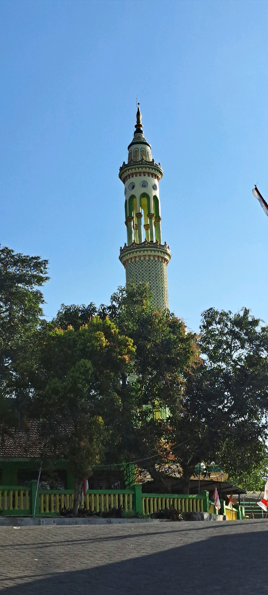 A tall, slender minaret, adorned with intricate patterns and painted in shades of white, green, and gold, stands prominently against a clear blue sky. The minaret is topped with a pointed golden spire. Lush green trees with dense foliage partially obscure the lower half of the minaret. In the foreground, a paved walkway or road with a brick pattern casts long shadows. Behind the trees, a building with a green facade and a tiled roof is visible, enclosed by a green and yellow striped fence. Indonesian flags are displayed on poles along the fence. The overall scene depicts a mosque minaret in the city of Sukoreno, Indonesia, likely on a sunny day.