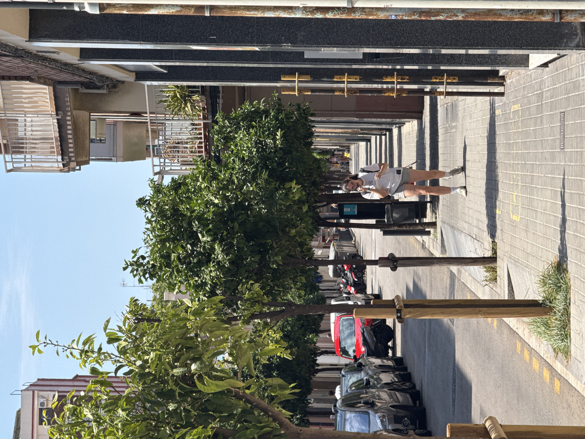 In L'Hospitalet de Llobregat, Spain, a woman is walking down a sidewalk lined with trees on a sunny day. The sidewalk is paved with light-colored square tiles and features yellow markings. To the left of the sidewalk, parked cars and motorcycles are visible on a street with a yellow dashed line marking the edge of the road. In the background, multi-story buildings with balconies and windows overlook the street. The woman, wearing a white t-shirt and grey shorts, is looking at her phone and has a backpack on her shoulders. She is walking towards the right side of the image. The scene depicts a typical urban street environment with pedestrian and vehicular activity.