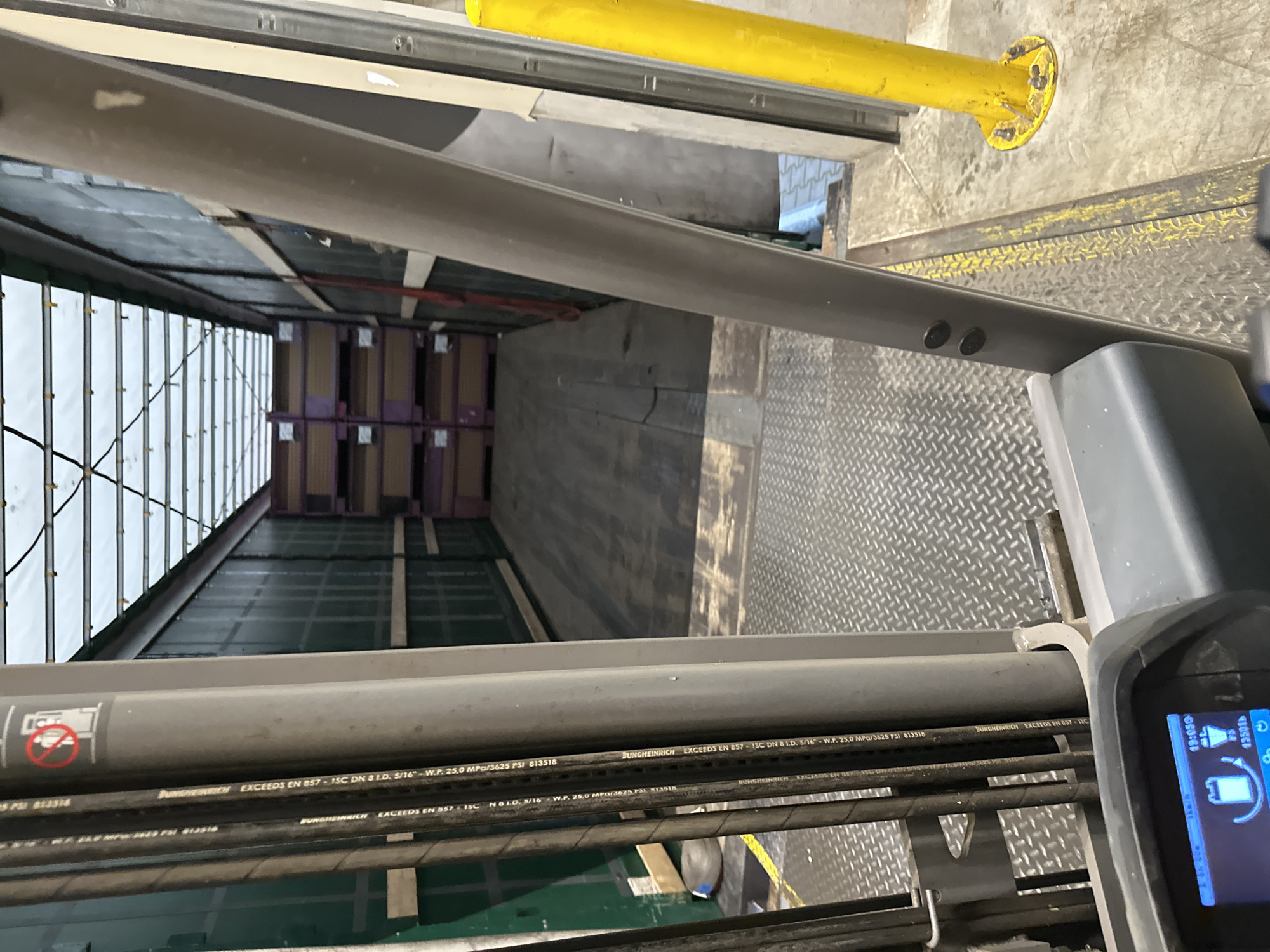 This media file captures a view from inside a warehouse or loading dock, looking down the length of a trailer or cargo bay filled with stacked boxes. The boxes appear to be on pallets and are predominantly purple and brown. The interior of the trailer is lined with metal and some white paneling.
In the foreground, there are several thick, grey hoses or cables running horizontally. Visible text on these hoses indicates they are "DUNGHEINRICH" products, specifically "EXCEEDS EN 857-1SC DN 8 I.D. 5/16" with a working pressure of "25.0 MPa/3625 PSI". Part of a handheld electronic device, possibly a scanner or control unit, is visible on the right side of the frame. The screen displays a time of "19:05" and some battery and speed indicators.
In the upper portion of the image, a bright yellow cylindrical object, likely a safety bumper or support, is mounted on a grey wall. The overall setting suggests an industrial or logistics environment. The lighting is consistent with an indoor, potentially artificial, light source. The specific location is provided as Brodce, Czechia. No people are visible in the frame. The image is oriented in a portrait aspect ratio.