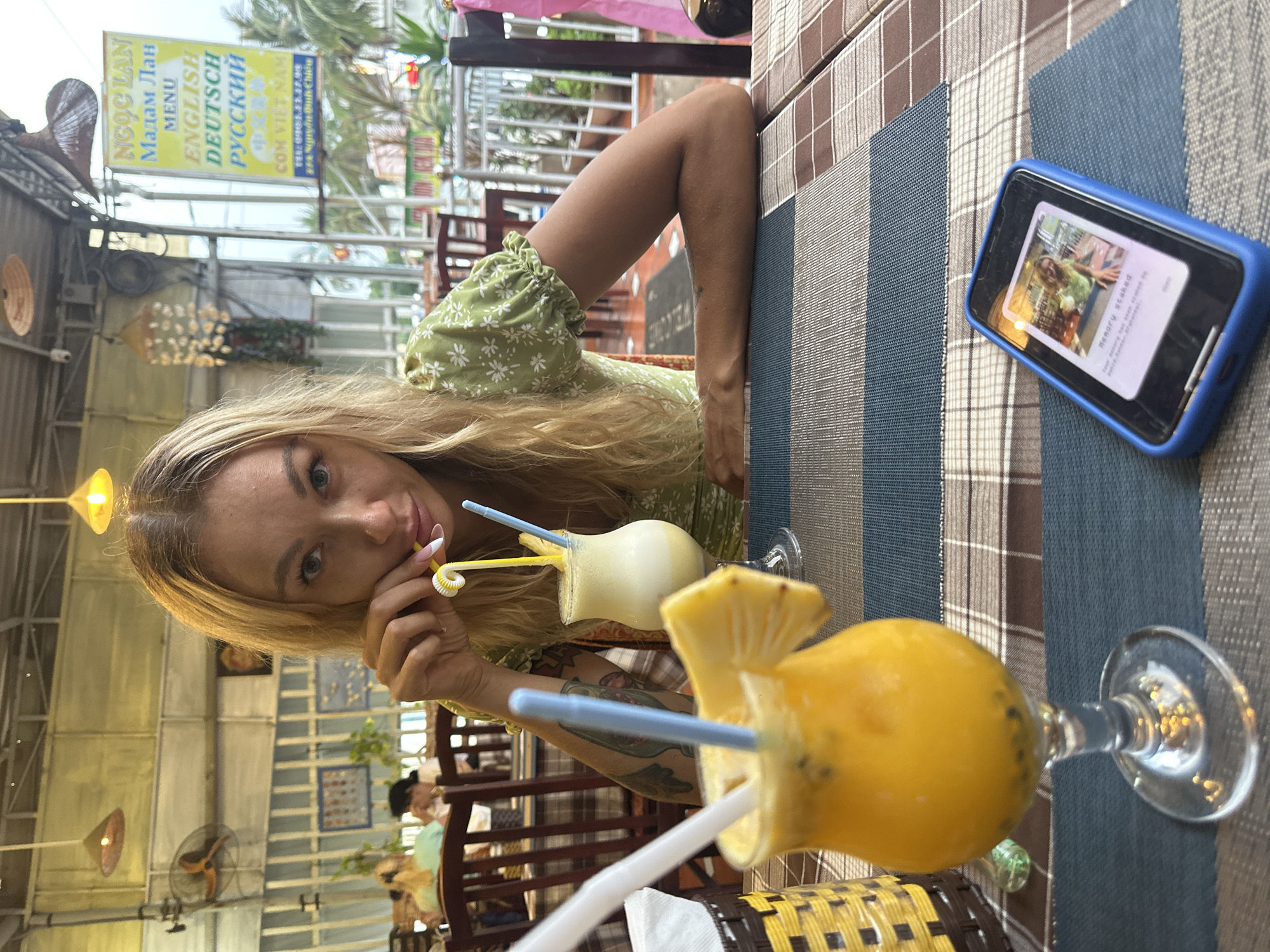 A woman is photographed at a table, likely in a restaurant in Phu Thuy Ward, Vietnam. She has fair skin, blonde hair, and is wearing a light green floral print dress. Two drinks, one yellow and one white, are in front of her, each with a straw. She is holding the straw of the white drink. A blue-cased phone is to the right of her. The table has a patterned covering. Outside, a sign reads "NGỌC LAN Мадам Лан MENU ENGLISH DEUTSCH РУССКИЙ COM VIET NAM," indicating the name and menu languages of the establishment. The surroundings suggest an open-air setting with other tables and patrons visible in the background.