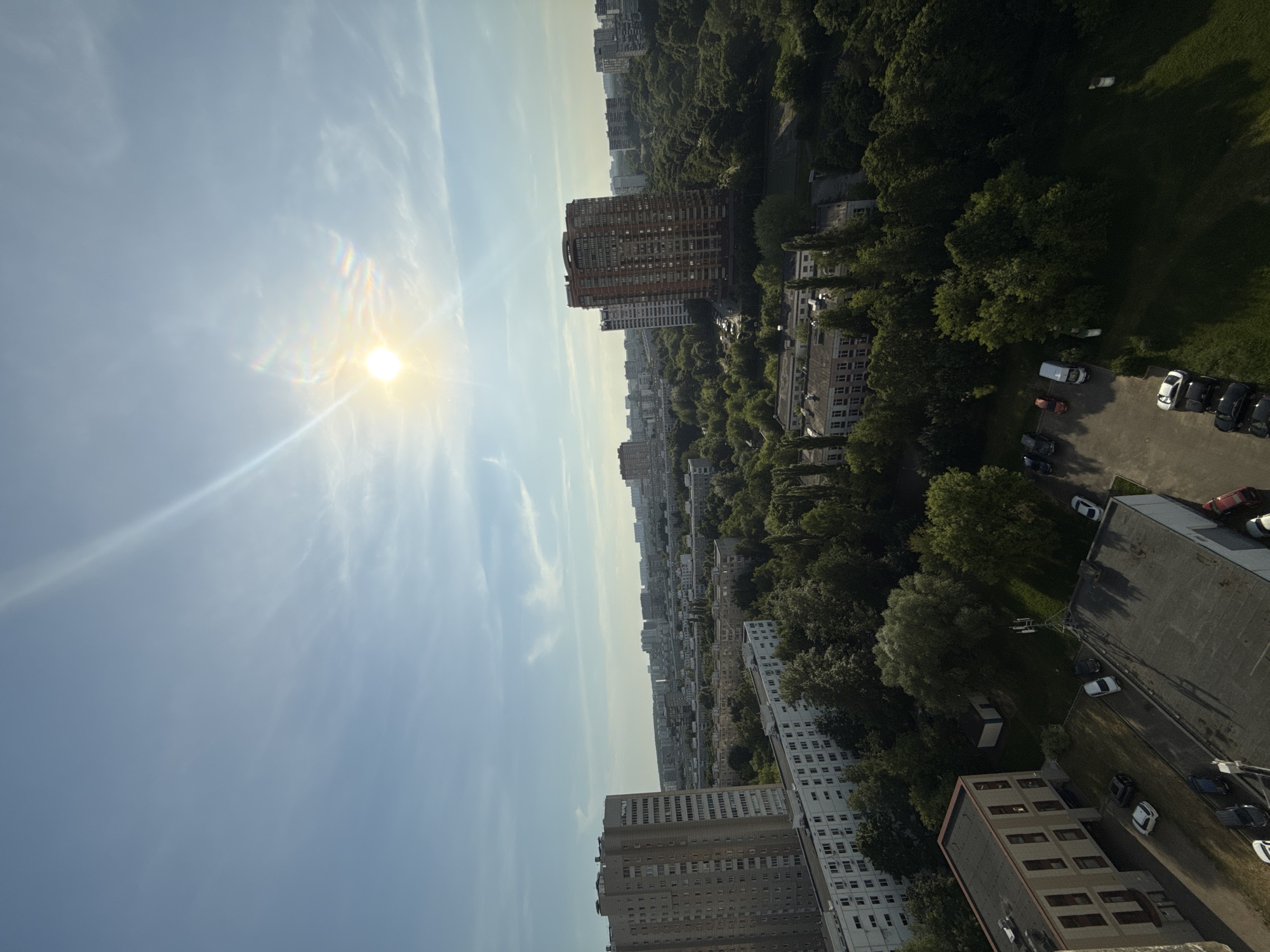 The image displays an elevated view of an urban landscape under a clear blue sky. The sun, positioned in the upper right quadrant, is bright and generates a prominent lens flare and sunbeam across the sky. Sparse, wispy clouds are visible.
The scene features numerous multi-story buildings, primarily residential, interspersed with extensive areas of mature green trees that suggest parks or heavily vegetated courtyards. On the left side of the frame, a tall, light-grey multi-story residential building with numerous windows occupies the mid-ground. Below it, a longer, lower-rise white residential building is visible. In the immediate lower-left foreground, a modern, light-colored building with a dark roof and rectangular windows fronts a paved parking area containing multiple parked cars.
On the right side, a tall, reddish-brown multi-story residential building stands prominently amidst green tree cover. Further into the background, a dense array of city buildings extends to the horizon. In the lower-center foreground, a small white shed-like structure is situated in a grassy area, alongside a cellular antenna mast. Additional parked vehicles are scattered across various paved surfaces.
No individuals are visible in the image, and no specific actions or interactions are depicted. The architectural characteristics of the residential buildings, including both older panel-type structures and newer multi-story complexes, alongside the extensive integration of green spaces within a dense urban fabric, are consistent with the city of Moscow, Russia. The bright sunlight and distinct shadows suggest the time of day is mid-day to late afternoon.