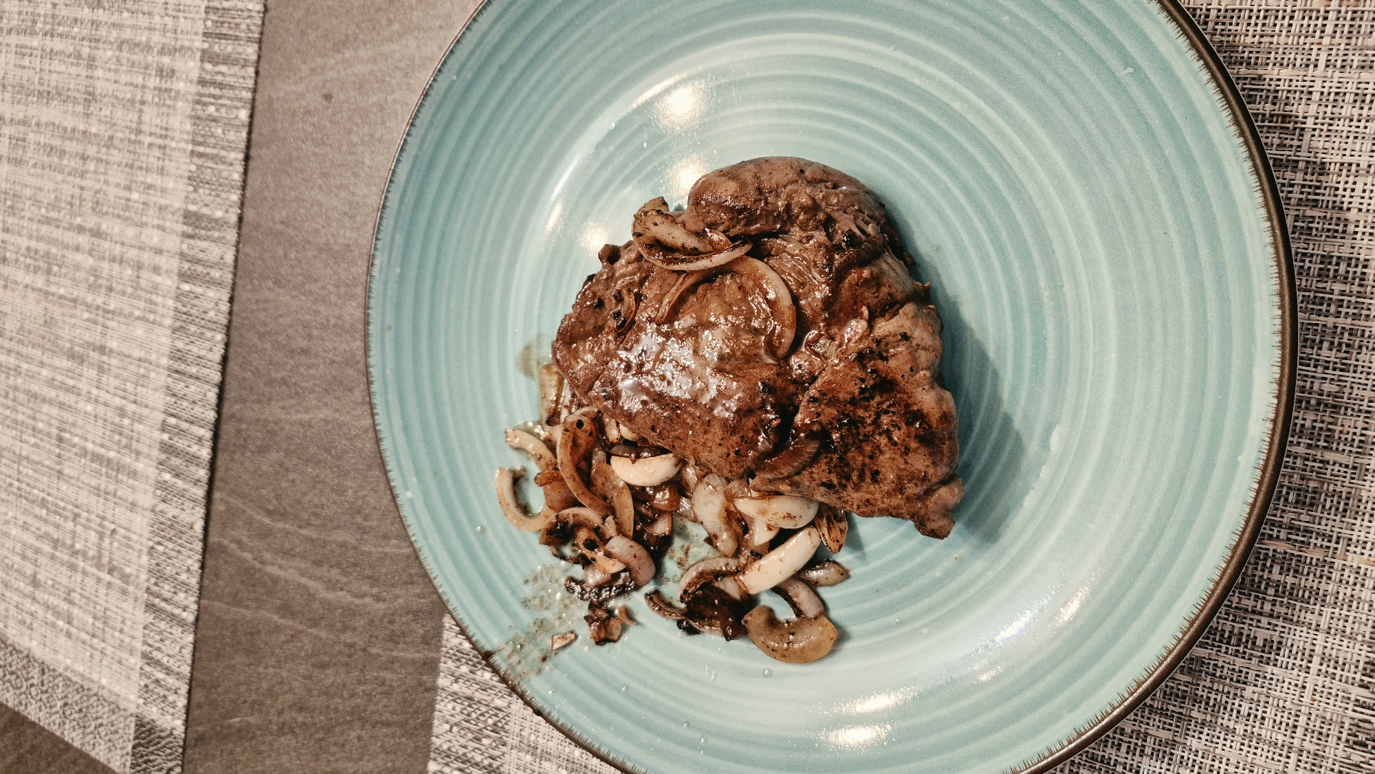 A close-up, high-angle shot features a cooked steak and sautéed onions on a teal-colored, ribbed plate. The steak, which appears to be a thick cut, is seared with grill marks and glistening with juices. The onions are thinly sliced and appear caramelized and slightly browned. The plate is positioned on a textured placemat, which is on top of a dark, speckled surface. The background shows a portion of a grey and white patterned placemat. The image appears to be taken indoors in a domestic setting. There is no indication of the city of Baden-Baden, Germany, in the image.