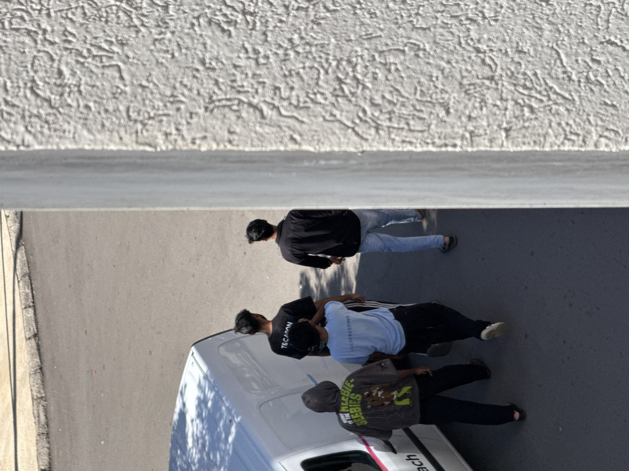 An overhead, high-angle view captures a street scene in Budva, Montenegro, with a white, heavily textured building wall partially obscuring the right side of the frame. Four males are present on a grey asphalt road. On the left, a white van, possibly a commercial or utility vehicle, is parked or stopped, with distinct shadows from a nearby tree cast upon its roof and side. Closest to the van on the left, a male in a dark grey hooded sweatshirt, bearing the text "NIGHT OF THE GEEBIES" and a cartoon character on its back, wears dark pants and sandals. To his right, another male wears a light blue t-shirt with "HOUSEKEEPING" printed across the back, dark athletic shorts or pants, and white sneakers. Slightly behind and to the right of the "HOUSEKEEPING" individual, a third male wears a black t-shirt with "T&CAPON" visible on the back, dark athletic pants with white stripes, and dark footwear. On the far right, partially obscured by the white building wall, a fourth male in a dark top and light blue jeans is visible, moving in the same general direction as the others. All individuals are seen from behind, facing away from the camera, suggesting movement forward along the street. The scene is brightly illuminated by direct sunlight, casting crisp shadows. A concrete curb runs along the edge of the asphalt road.