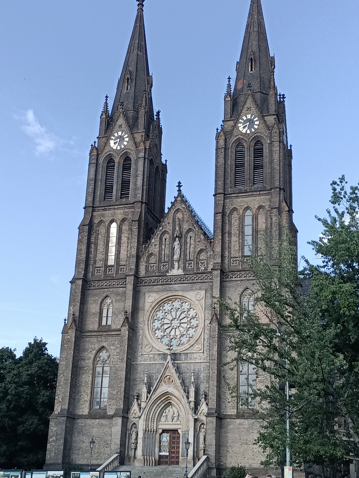 This is a vertical, eye-level shot of the Basilica of St. Peter and St. Paul in Prague, Czechia. The imposing Gothic Revival facade dominates the frame, with two tall spires reaching towards a clear blue sky. The spires are adorned with crosses and intricate stonework. Below the spires, large clocks are visible on each tower, though their hands are difficult to discern. A prominent rose window, a masterpiece of stained glass artistry, is centered on the facade, surrounded by arched windows and decorative carvings. At the base of the church, a grand entrance with double wooden doors and a stone staircase is visible. Statues of saints can be seen in niches on either side of the entrance. Lush green trees frame the right side of the church, partially obscuring the building and suggesting an outdoor setting. The lighting indicates it is daytime, likely late afternoon or early evening, given the warm tones on the stone. The scene appears peaceful, with no discernible activity except for a few people near the bottom of the frame, suggesting they are visitors or passersby. The architecture and sky suggest a pleasant, clear day.