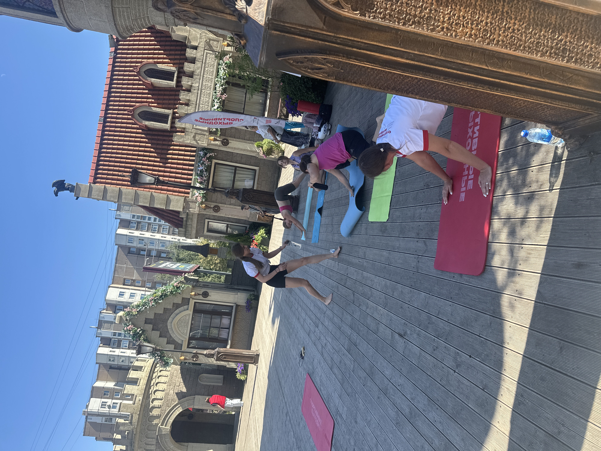 The image shows a group of women practicing yoga on a wooden deck in front of a building with a fairy-tale aesthetic. The building features whimsical architectural elements like a turret with an eagle statue, arched windows, and decorative floral arrangements. In the background, modern apartment buildings are visible, suggesting an urban setting. The weather appears to be sunny with a clear blue sky. One woman in the foreground is in a push-up position on a red yoga mat, wearing a white t-shirt and black shorts. Several other women are positioned on yoga mats in various yoga poses. A water bottle rests on the deck near the woman in the foreground. The scene suggests an outdoor yoga class or event. The specific location is in Moscow, Russia, at a place called "Skazka" (Fairy Tale), which is an amusement park.