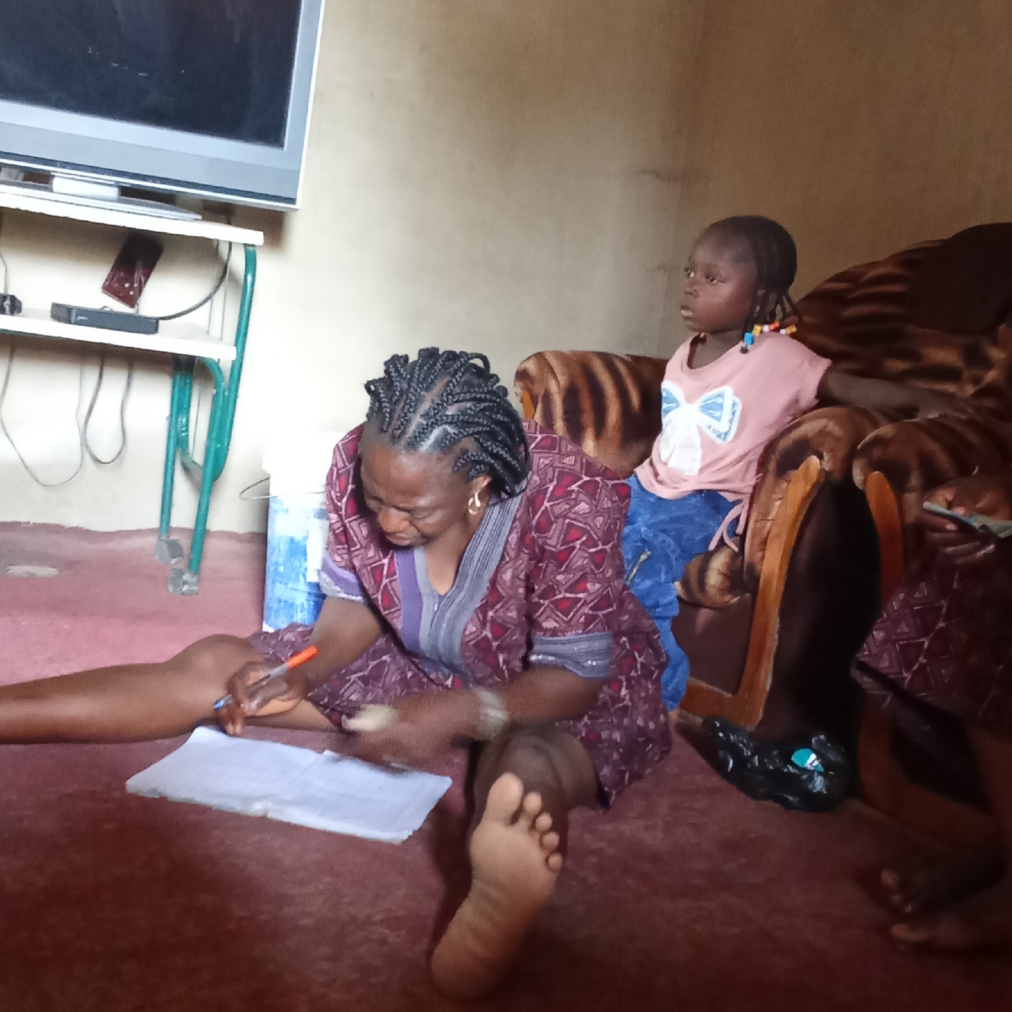 A woman with braided hair, wearing a patterned dress, sits on the floor writing on a notepad with a pen. Her bare feet are visible. To her right, a young girl with beaded braids sits on a brown patterned couch, looking towards the left. A television sits on a white stand with green legs in the background. The setting appears to be a simple room with off-white walls and a maroon-colored floor. A bucket is partially visible to the woman’s left. A black plastic bag is near the couch. The location is Akwanga, Nigeria.