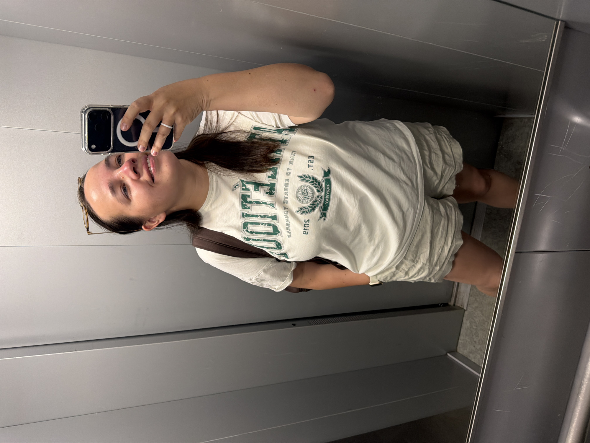 A woman is taking a selfie in an elevator. She is wearing a white t-shirt with green lettering and khaki shorts. She has a brown bag slung over her shoulder and eyeglasses pushed up on her head. The elevator has gray walls. The location of the elevator in the city of Chaikovsky, Russia, cannot be determined from the image.