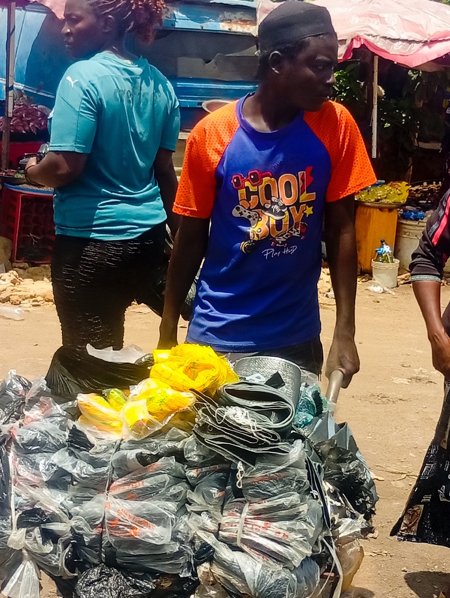 The image depicts a street scene in Bukuru, Nigeria, on a sunny day. Two people are the primary subjects. On the left, a woman with braided hair wears a teal t-shirt and black, textured pants. She is facing left and appears to be engaged in conversation or observing something.
To her right, a man wearing a black skullcap and an orange and blue t-shirt stands behind a large pile of goods. The t-shirt has graphics on it, including the words "COOL BOY" and "Play H₂O". He is holding onto a metal rod, suggesting he is pushing or maneuvering the cart of goods.
The objects in the foreground are a large assortment of items wrapped in plastic bags, primarily black and some yellow. They appear to be wares for sale, possibly textiles or similar goods, neatly bundled and stacked on a wheeled cart or trolley. Yellow plastic bags are also visible among the merchandise.
In the background, elements suggest a market or roadside stall environment. There is a blue structure, possibly a vehicle or stall, and a pink and white awning. The ground is sandy and dusty. The bright sunlight indicates it is daytime. The overall atmosphere seems to be that of a busy marketplace.