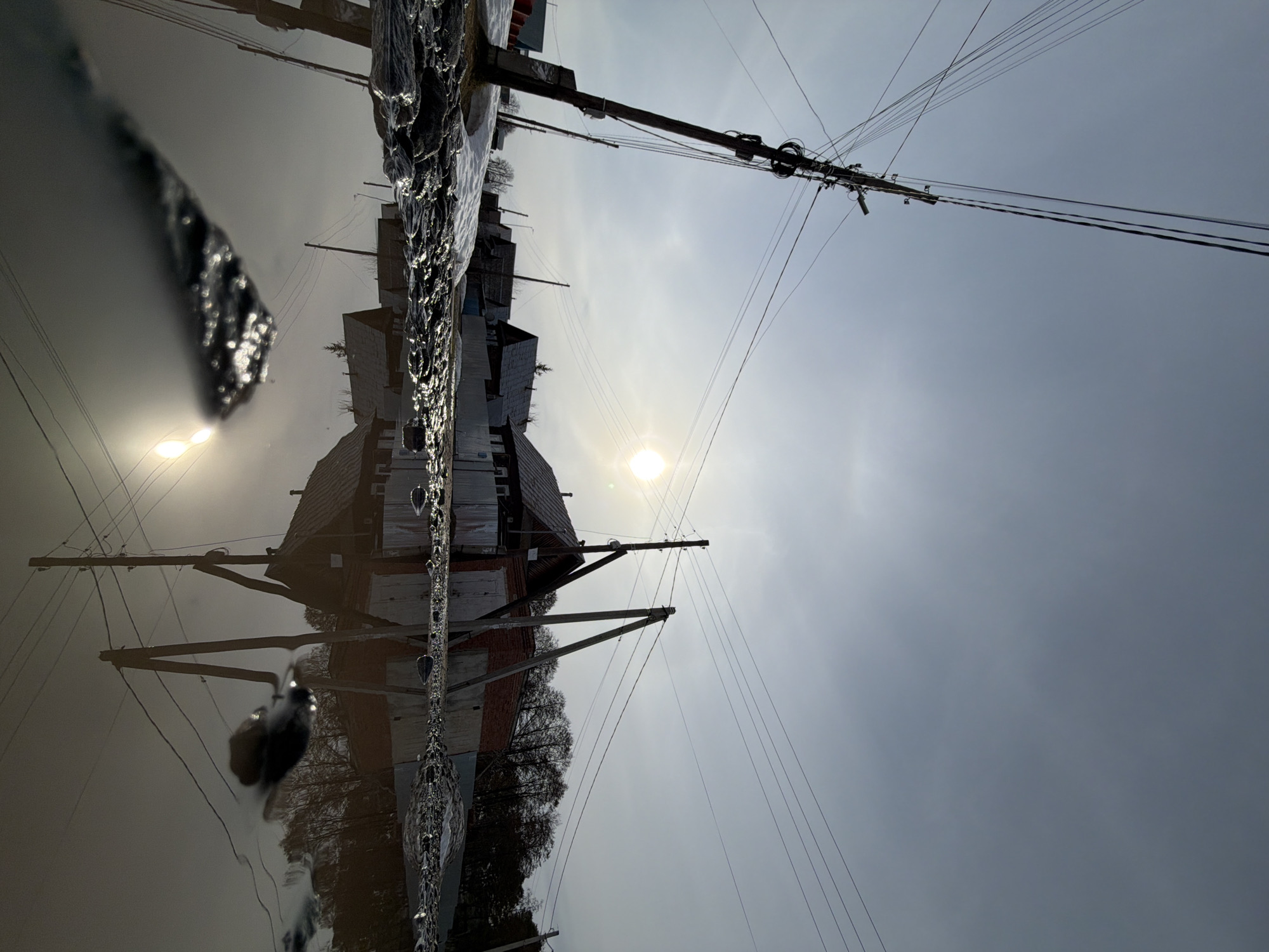The image appears to be a reflection of buildings and power lines in a body of water. The buildings are a mix of brick and wood construction, with a prominent gable roof. Electrical poles and lines crisscross the sky. The sun is visible, casting a bright light on the scene. The water reflects the buildings and sky, creating a mirrored effect. In the foreground there is a chain link fence covered in ice. Based on the city context provided, the location is likely in Chaikovsky, Russia.