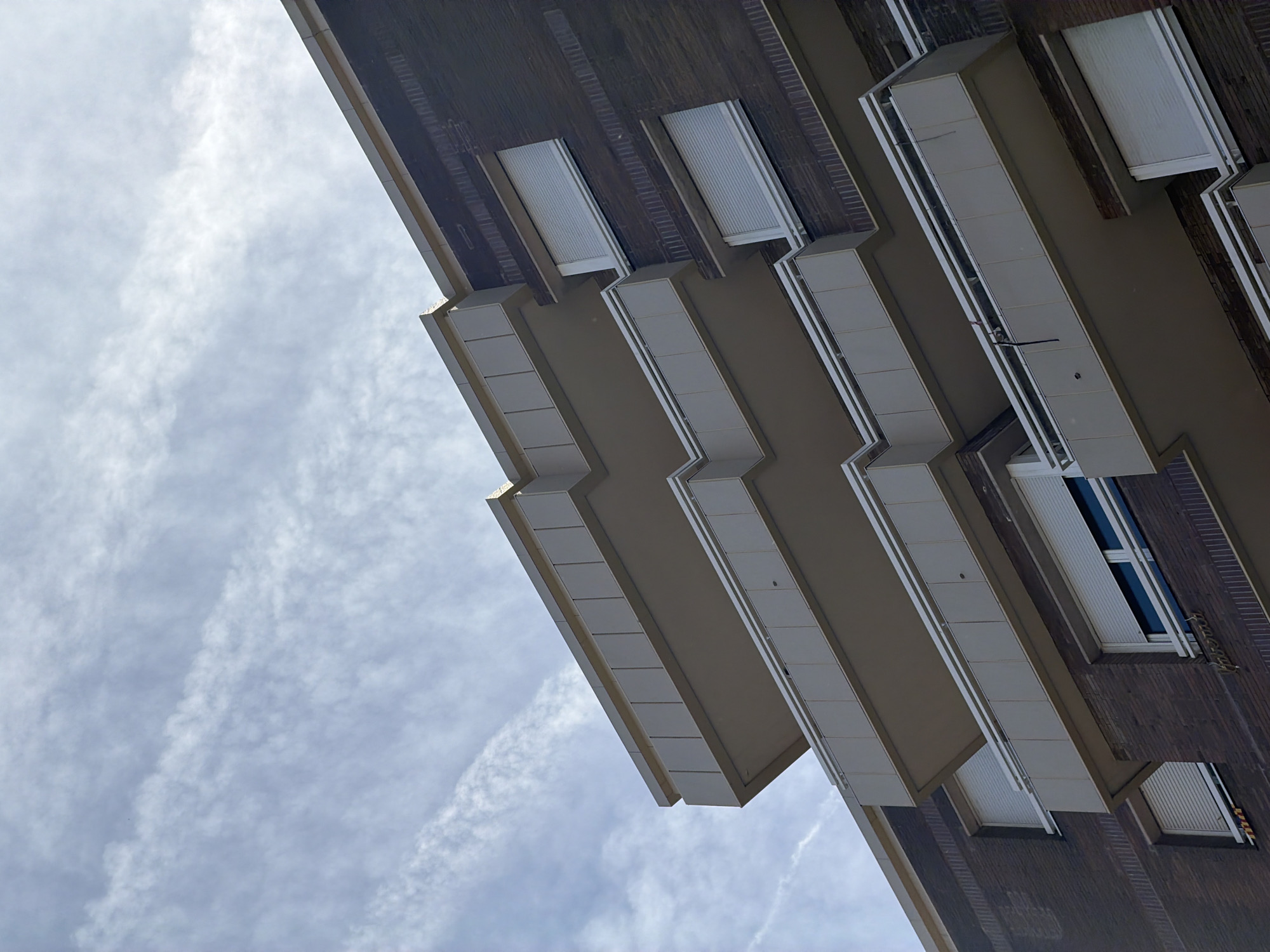 The image shows a low-angle view of a modern apartment building with multiple balconies against a cloudy sky. The building facade is constructed with dark brown brickwork, and the balconies are cantilevered with light-colored, angular detailing. White roller shutters are visible behind the balcony railings. The sky is filled with thin, wispy clouds, suggesting an overcast or partly cloudy day. No people or other significant activity are depicted in the scene. The architectural style suggests a contemporary urban setting.