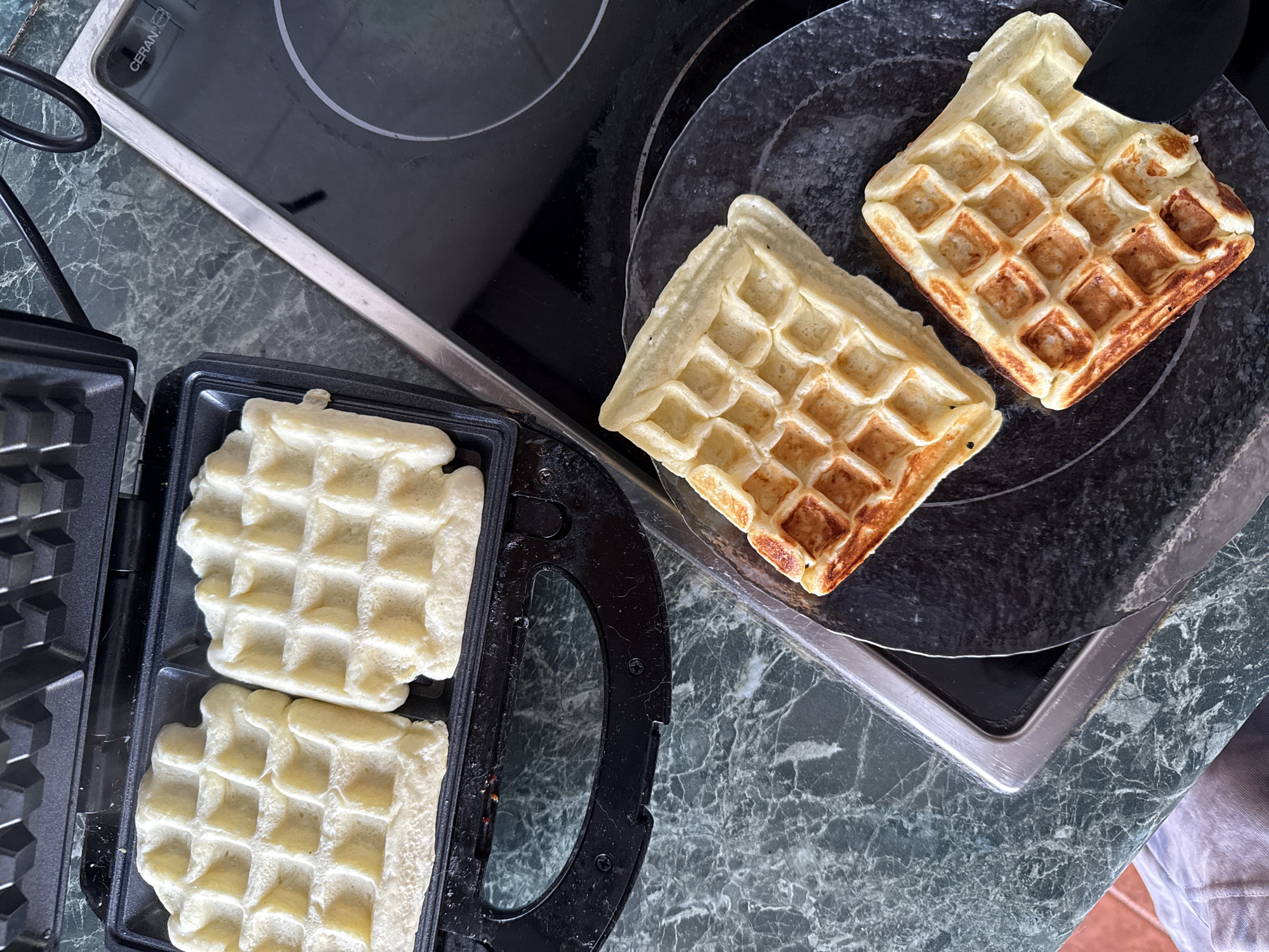 On a countertop with a green marble pattern, a waffle maker is open, revealing two uncooked waffles. To the right, two cooked waffles sit on a black circular plate placed on a black stovetop. The scene takes place in an indoor kitchen setting. There is no evidence to suggest the image was taken in Krasnoyarsk, Russia. No people are visible.