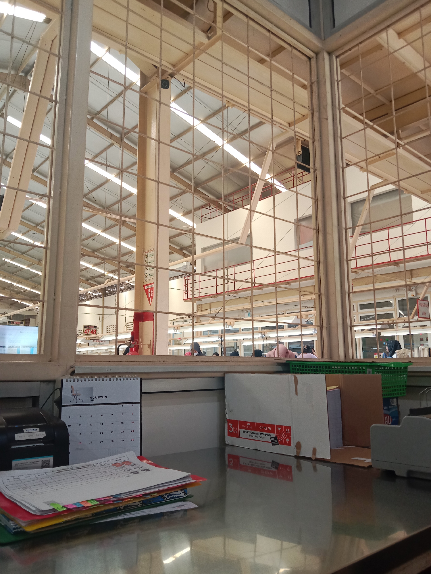 The image depicts an indoor view of a factory or warehouse environment, likely in Semarang, Indonesia. The foreground is dominated by a metal desk with various items. To the left, there is a black Xprinter, a device commonly used for printing receipts or labels. Beside it, a stack of colorful folders and papers with sticky tabs are piled on the desk. A spiral-bound desk calendar for "AGUSTUS 2020" is visible, showing a grid of dates and an image of an office chair.
Further back, through a large, metal-grated window or partition, the interior of the facility is visible. The space appears to be a large, open area with a high, industrial ceiling supported by metal beams and trusses, allowing natural light to filter through translucent panels. There are multiple levels within the structure, indicated by elevated walkways and railings. Some workers, partially obscured by the grate, can be seen in the distance, engaged in various tasks. One individual wears a pink shirt, and another wears a headscarf. Several boxes, including a "3 PLY KRAFT" cardboard box and a green plastic basket, are present on a counter or surface in the middle ground. The overall impression is a functional, operational industrial space.