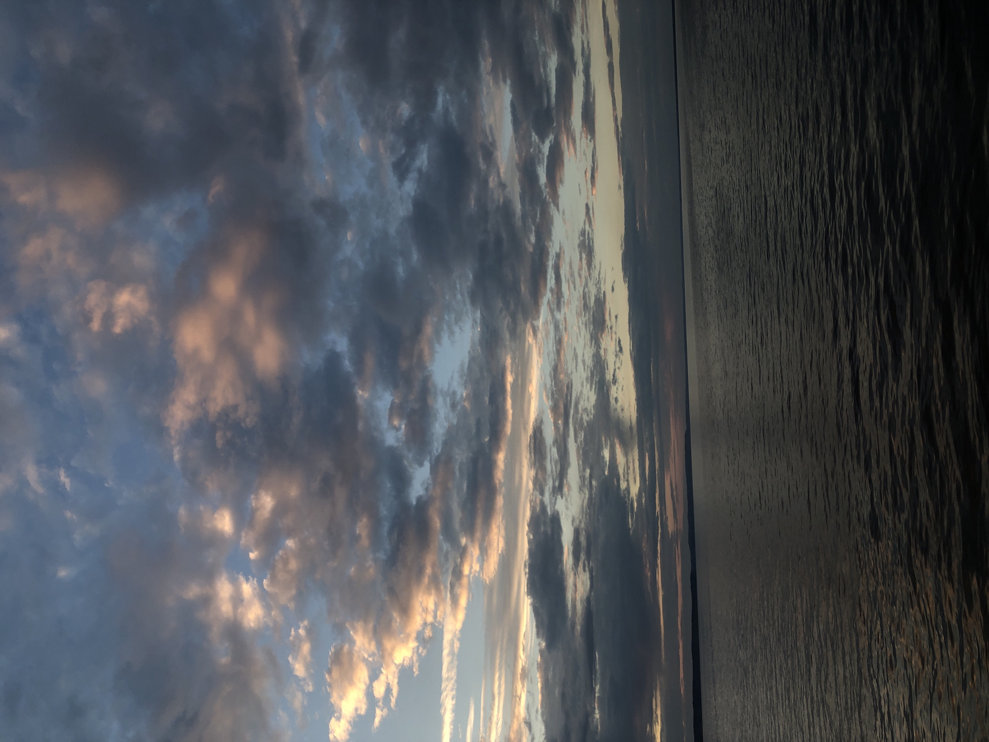 The image captures a wide expanse of water, likely a reservoir or a large river, at dusk. The water's surface is rippled, reflecting the muted colors of the sky. A dark, elongated landmass is visible on the horizon, suggesting the presence of hills or a distant shoreline. The sky is filled with dramatic clouds, tinged with shades of gray, blue, and hints of pink and orange from the setting sun. The overall atmosphere is serene and somewhat melancholic. This scene is consistent with views of the Kama River reservoir in Chaikovsky, Russia, known for its picturesque sunsets over the water and the low hills on the opposite bank.