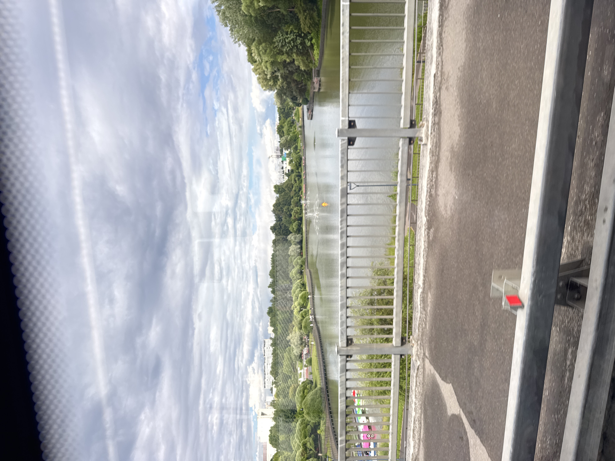 The image shows a canal or river with a walkway and trees lining its banks. A metal railing separates the walkway from a paved surface, possibly a road or bridge. In the distance, buildings and more trees are visible. The sky is cloudy with patches of blue. The scene appears to be a park or recreational area. There are no discernible people or specific objects of interest, besides what appears to be a small red marker attached to the railing. The image is taken from a slightly elevated perspective, looking across the water and towards the opposite bank. The overall impression is one of a tranquil, green environment. The orientation of the image is vertical.