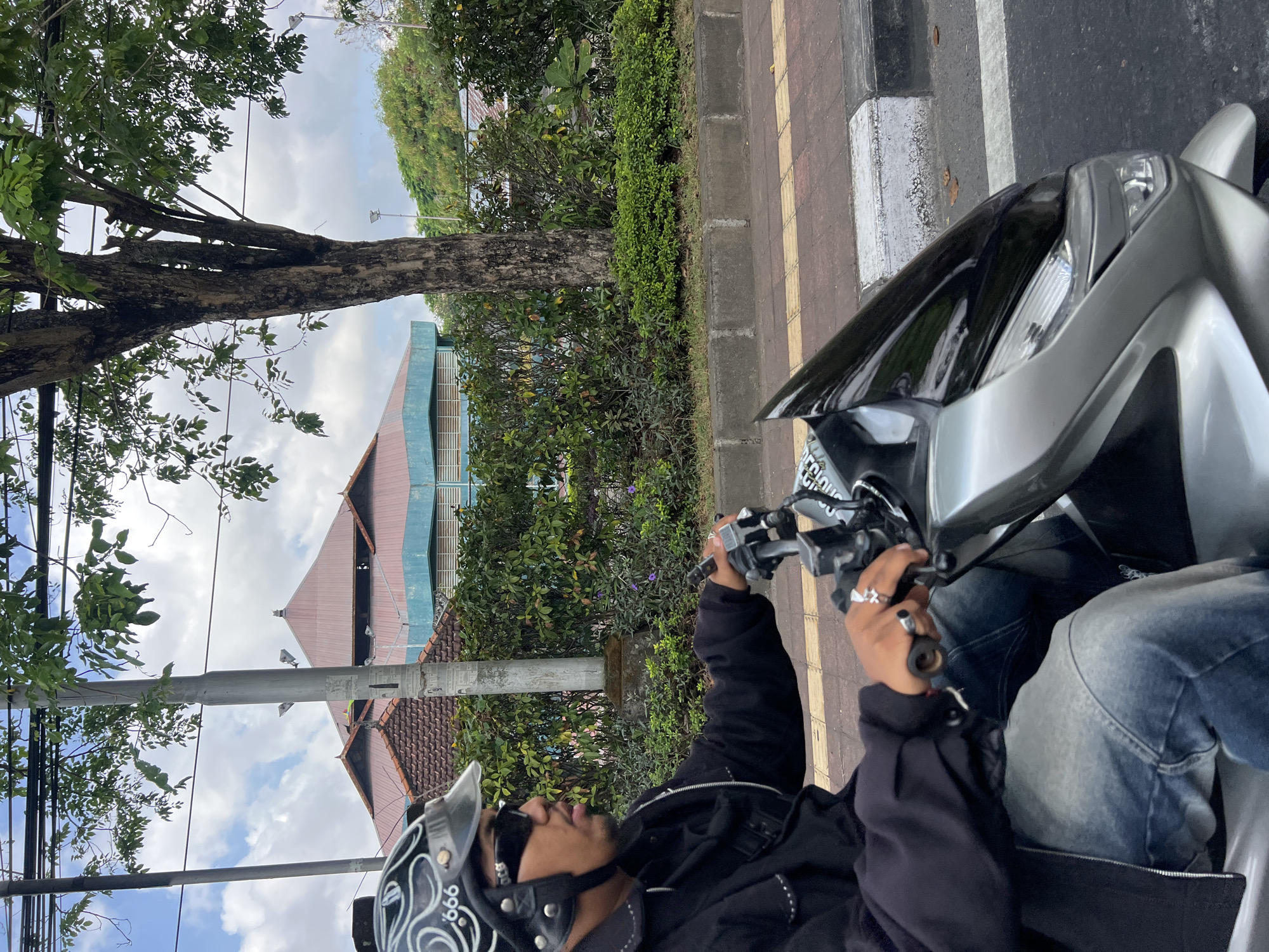 A man wearing a black jacket, blue jeans, sunglasses, and a black helmet with white designs is riding a silver scooter in Denpasar, Indonesia. He is looking to the left while holding onto the scooter's handlebars with his right hand. The scooter is on a road next to a sidewalk with some greenery and a building with a red roof in the background. The sky is cloudy.