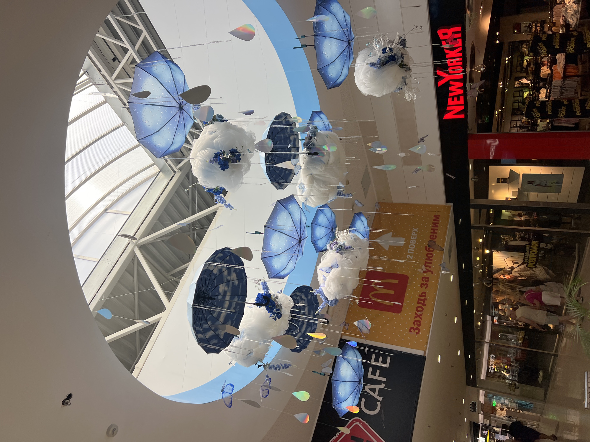 The image shows an indoor shopping mall, likely in Fontanka, Ukraine, decorated with a whimsical, sky-themed display. Hanging from the ceiling are numerous blue umbrellas, some appearing to have a cloud pattern, interspersed with fluffy white cloud-like decorations adorned with blue flowers and small butterflies. Dangling from strings are iridescent, teardrop-shaped ornaments that mimic raindrops, as well as silver beads. Below this overhead display, signs for "CAFE" and "McDonald's" are visible, with the latter featuring Cyrillic text. Further to the right, a storefront with a prominent red "NEW YORKER" logo indicates another retail establishment. In the lower portion of the image, people can be seen walking through the mall, some browsing in shops. The overall atmosphere is brightly lit, suggesting a public indoor space.