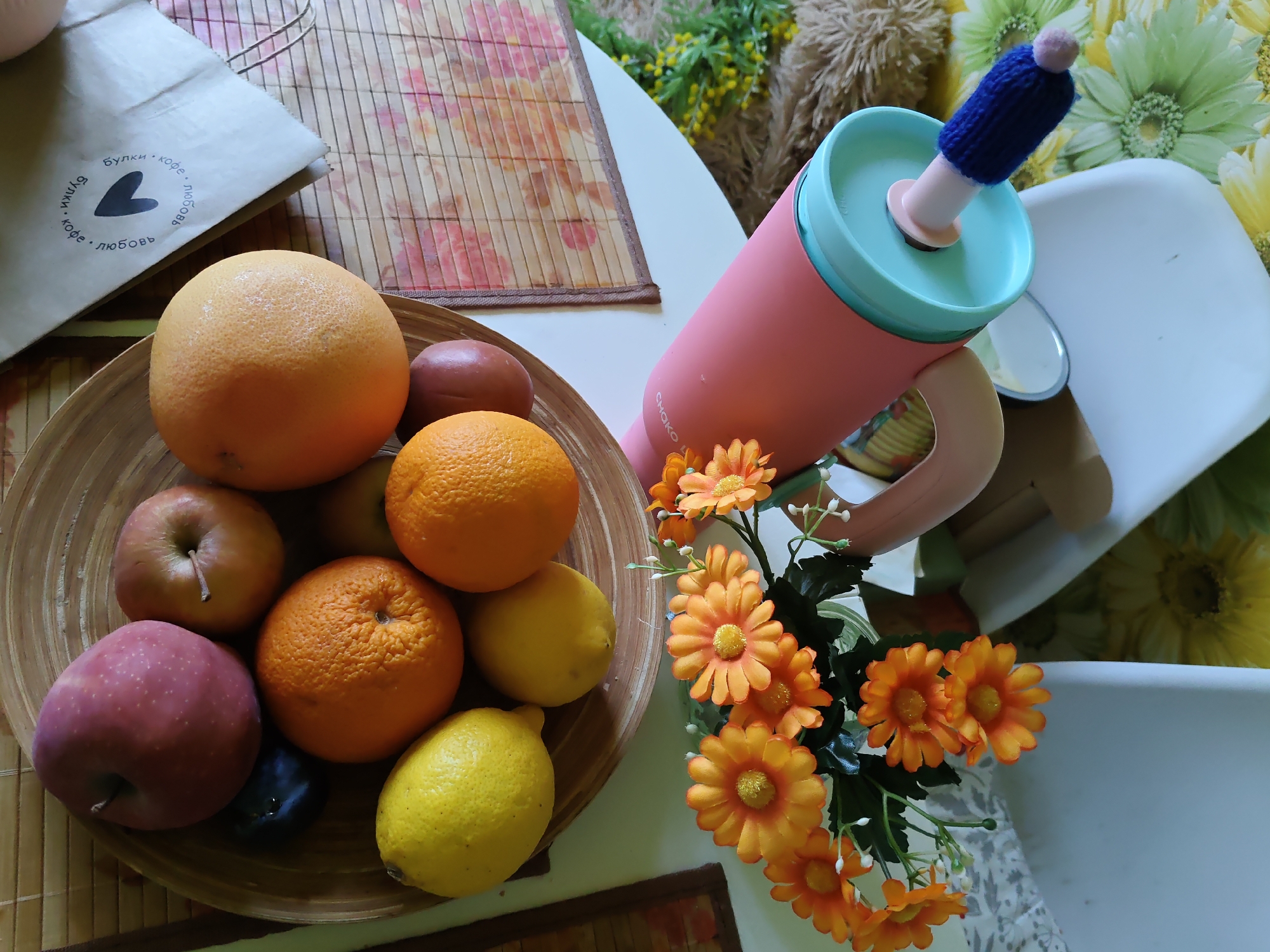The image depicts a still life arrangement of fruits and a thermal mug on a table. In the foreground, a wooden bowl contains several fruits, including two oranges, two lemons, two apples, and a plum. Behind the bowl, a pink thermal mug with a teal lid stands upright. The mug has a blue knitted cover on its straw. To the left of the bowl is a brown paper bag with text and a heart logo. To the right of the mug are orange artificial flowers. The background shows a table covered with a woven placemat, a portion of a chair, and floral fabric. The image is captured from a top-down perspective. The text on the paper bag includes Cyrillic characters. This image was taken in the city of Rostov-on-Don, Russia.