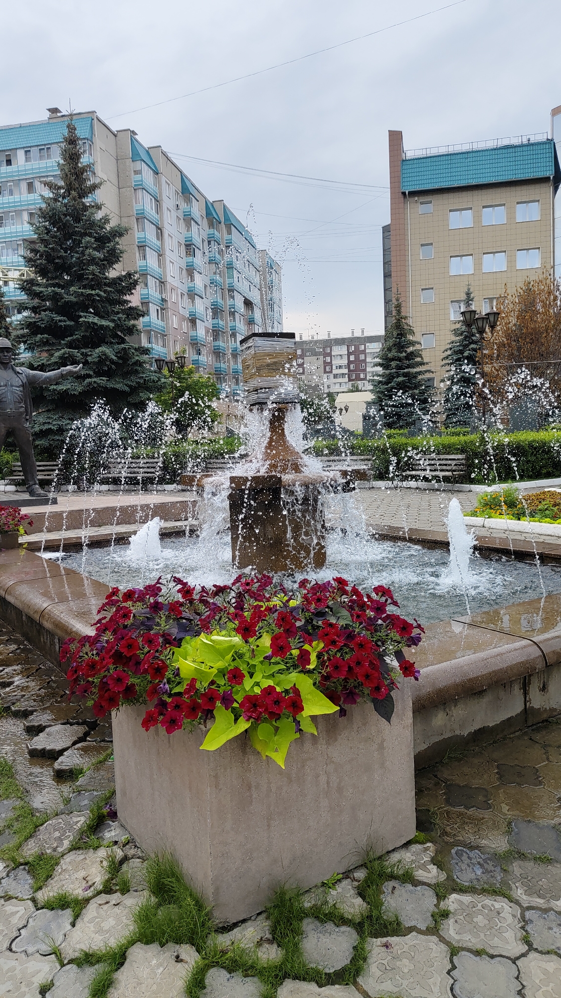The image depicts a public square in Krasnoyarsk, Russia, featuring a multi-tiered fountain with water spraying upwards. In the foreground, a large concrete planter overflows with vibrant red petunias and bright green foliage. The fountain itself is made of stone and has multiple streams of water. Behind the fountain, a bronze statue of a man in work clothes stands to the left, pointing forward. Several evergreen trees are visible, along with benches and paved walkways. In the background, a row of modern, multi-story apartment buildings with light blue trim lines the street, interspersed with other office buildings and trees. The sky is overcast.