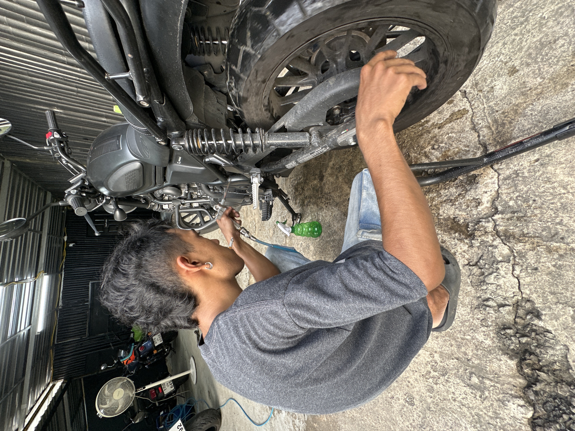 A young man with a mullet haircut and a silver hoop earring is crouching down to clean a black motorcycle in what appears to be a garage or workshop located in Karon, Thailand. He is wearing a grey t-shirt and light blue jeans, and black slides on his feet. The man is holding a spray bottle with green liquid in his left hand and is aiming a spray nozzle from a hose towards the motorcycle's rear wheel. The motorcycle is dark-colored with chrome accents and has a visible rear suspension system. In the background, there are other motorcycles, a large white fan, and corrugated metal walls. The ground is concrete with visible cracks and some water or cleaning solution spilled on it. The lighting suggests it is daytime, with artificial lights visible on the ceiling.