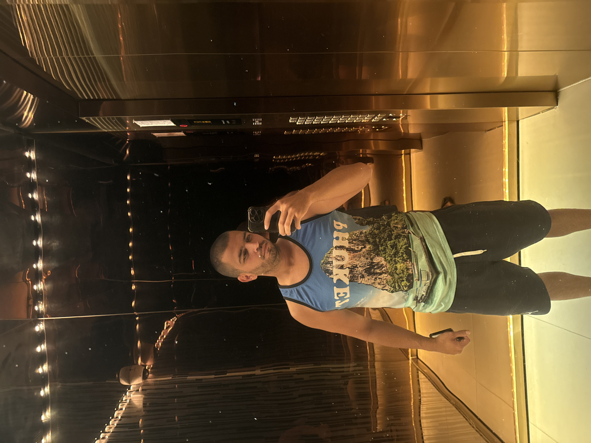 A man is captured in a selfie taken in a highly reflective, opulent interior, likely an elevator, in Kuala Lumpur, Malaysia. The image is oriented with the man standing vertically in the center. He is fair-skinned, bald, and has a short beard and mustache, looking directly at the camera with a neutral expression. He is wearing a blue and light green sleeveless tank top with a printed design of a mountain range, over which the mirrored text "BROOKLYN" is visible. He is also wearing black shorts.
The man holds a smartphone in his right hand to take the photo. The surroundings are characterized by dark, polished, reflective surfaces, possibly bronze or mirrored metal, on the walls and ceiling, creating numerous reflections of the man and the environment. Warm, golden-toned lighting emanates from unseen sources, casting linear reflections across the ceiling and walls.
In the reflections, a control panel resembling an elevator panel is visible, featuring a grid of buttons and some indistinct mirrored text, which includes phrases like "EMERGENCY CALL," "DOOR OPEN," "DOOR CLOSE," and various numbers. The floor appears to be a lighter, reflective material, showing reflections of the man's lower body and the ambient light. The overall impression is one of modern, upscale interior design.