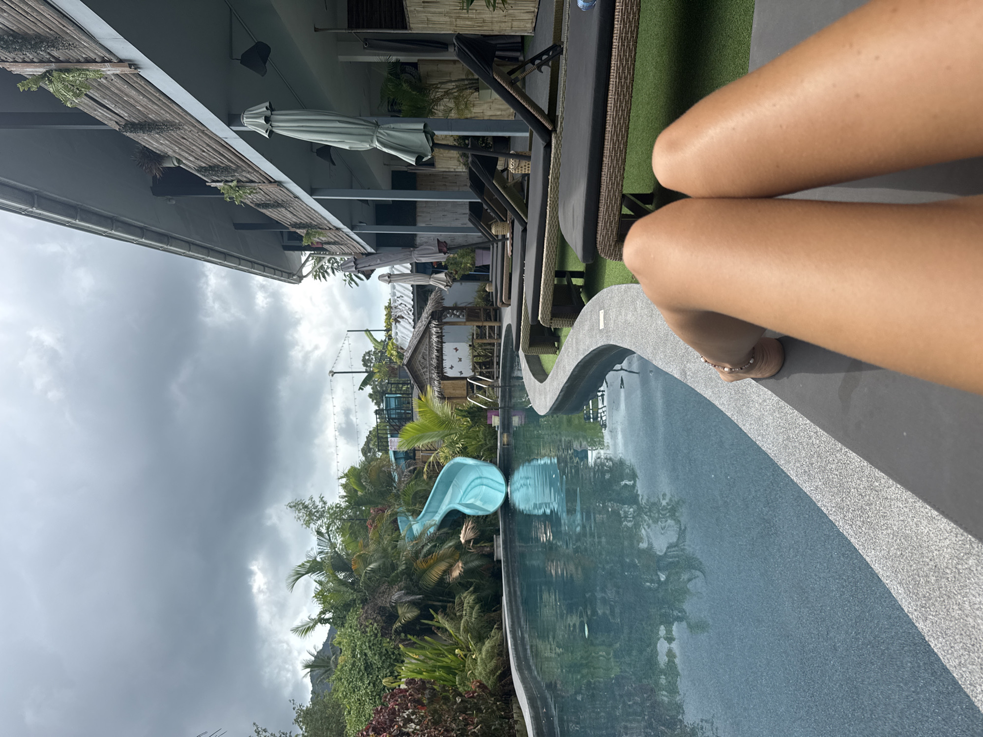A wide-angle, slightly rotated, outdoor shot shows a woman’s legs on a lounge chair next to a pool at a resort in Baan Mae Nam, Thailand. The woman’s legs are light brown and positioned vertically in the right foreground. She wears a tan sandal and an anklet with white beads. A curved pool edge of grey speckled stone runs from the foreground. The pool reflects the surrounding foliage and cloudy sky. Beyond the pool, tropical plants and trees cover the area, including a bright blue water slide. Further back, buildings with thatched roofs are visible. Lounge chairs line the pool. The sky is overcast with grey clouds. Building exteriors with green vegetation and awnings can be seen on the left.