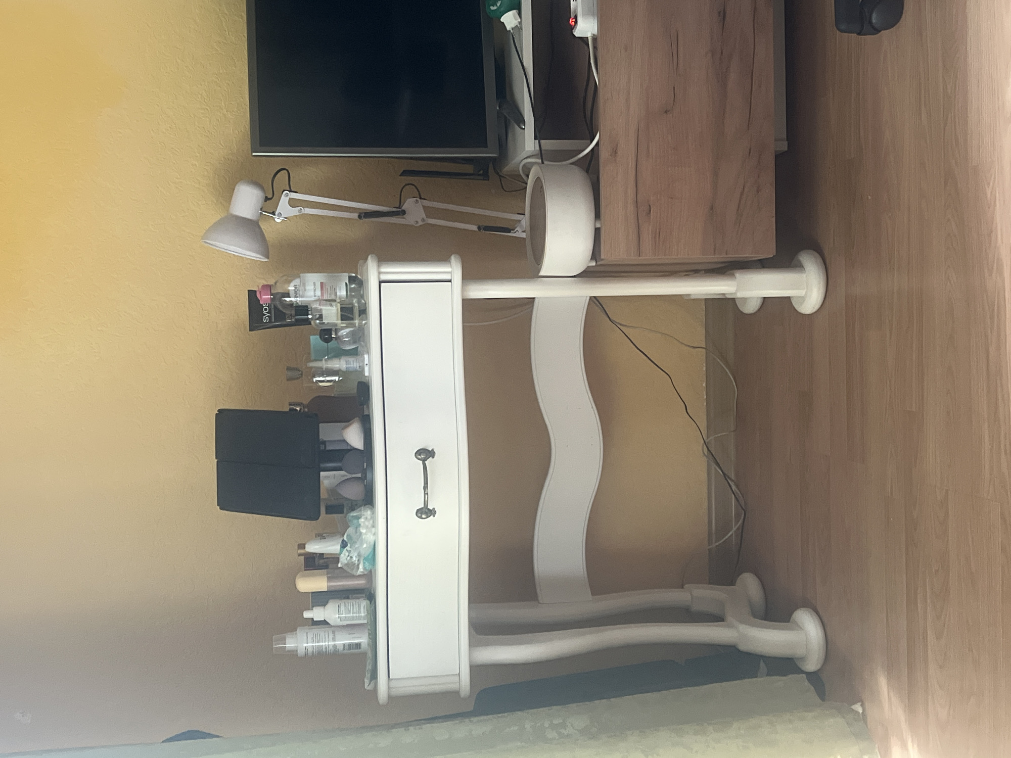 A white vanity table with a single drawer and a metal pull is positioned against a yellow textured wall. Various cosmetics and toiletries are arranged on top of the vanity, including bottles, jars, cotton swabs, and makeup sponges. A black foldable mirror is also present. To the right of the vanity, a modern desk lamp with a white shade is attached to a white pole. Next to the lamp, a television screen is visible on a wooden cabinet. A white, bowl-shaped object with a brown top sits on the cabinet, connected by a white wire. Electrical cords and a power strip are present on the floor and the cabinet. The floor is made of light-colored wood planks. A pale green curtain hangs on the left side of the frame. The scene is indoors, likely a bedroom or dressing area. There are no people or animals visible in the image. The location is not specified.