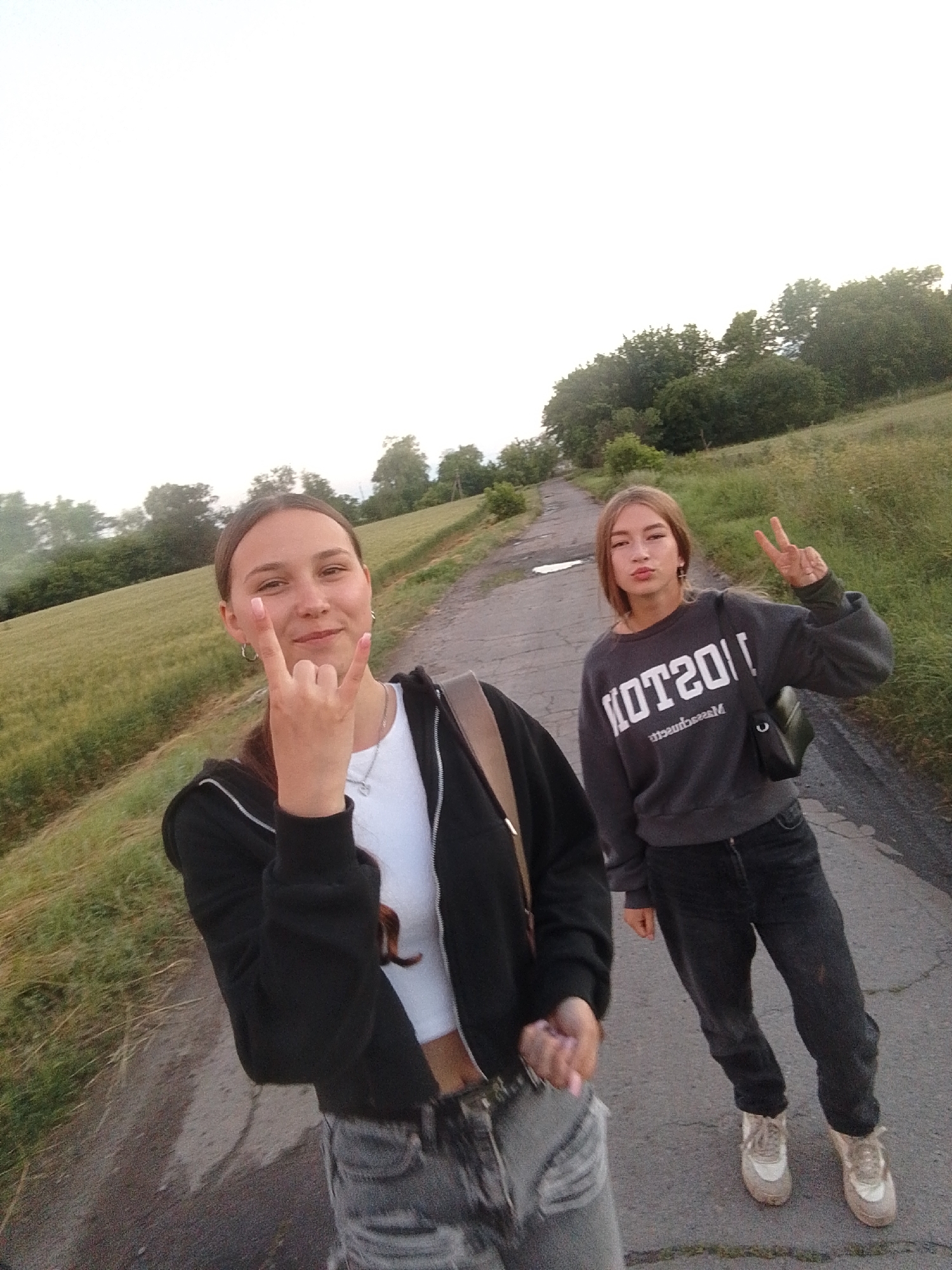 Two young women are posing for a selfie on a paved path in a rural, grassy area. The woman in the foreground on the left is wearing a black zip-up hoodie over a white top and grey distressed jeans. She has her right hand raised, forming a rock-and-roll gesture. The woman on the right is wearing a dark grey sweatshirt with "BOSTON" printed in white across the front, black jeans, and white sneakers. She is holding her left hand up in a peace sign. The background shows a gentle slope of grassy hills with scattered trees. The sky is bright white, suggesting it is either early morning or late afternoon. The location is identified as being in Ustyvytsia, Ukraine.