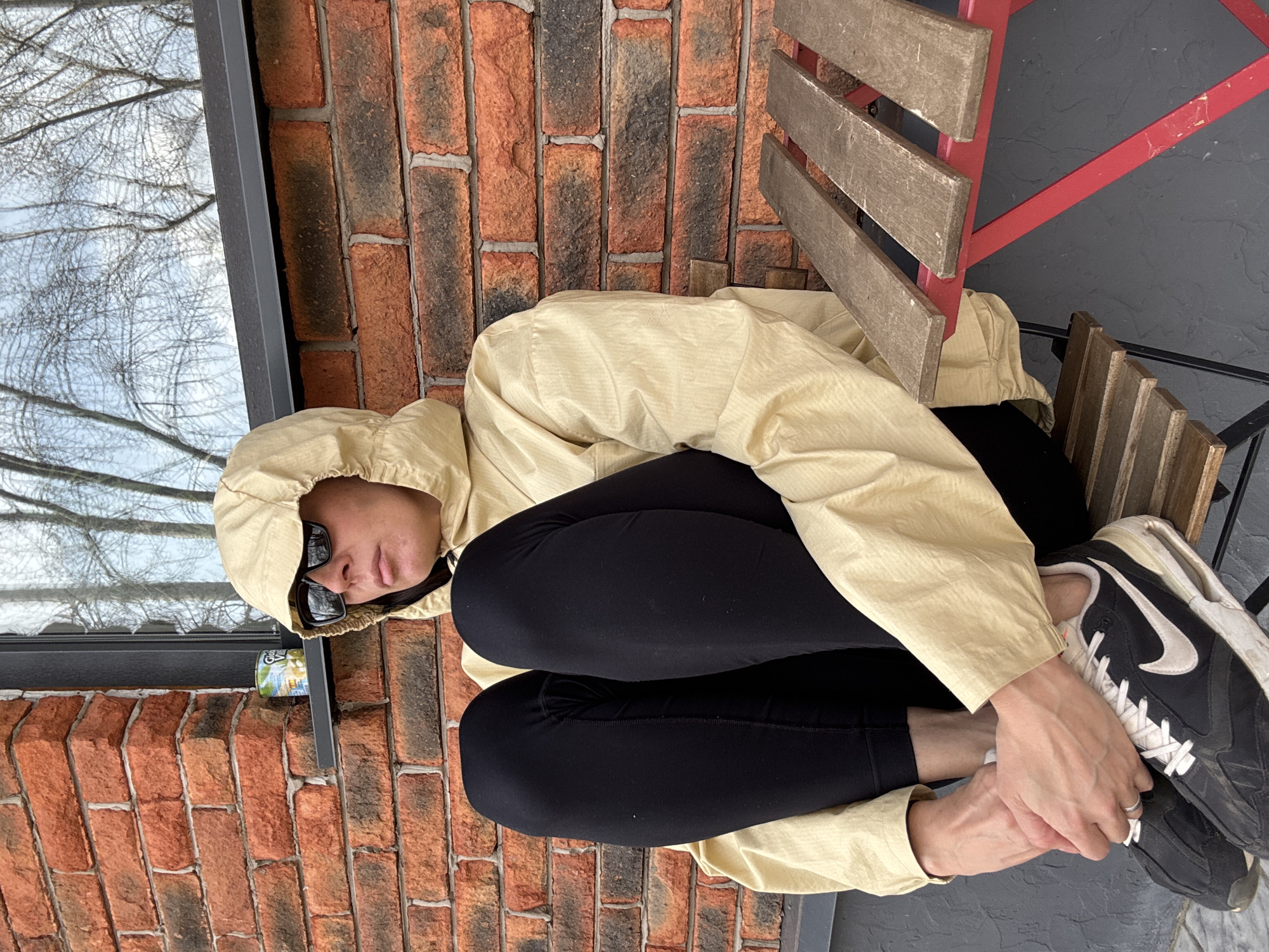 An individual is depicted sitting outdoors in Chaikovsky, Russia, with their body oriented towards the viewer's left. They are wearing a light beige or yellow hooded jacket pulled over their head, black leggings, and black and white Nike athletic shoes. Their hands are clasped over their right foot, which is resting on their left knee. Dark sunglasses obscure their eyes.
The person is positioned against a background featuring a textured brick wall to their left and behind them. To the left of the person is a window reflecting the bare branches of trees, suggesting either late autumn, winter, or early spring. On a dark ledge beneath the window, a small can with partially visible text, possibly "Yogurt," is placed. To the right of the individual, a chair with wooden slats and a red metal frame is visible. The ground appears to be a dark, possibly concrete or tiled surface. The lighting suggests daytime, likely overcast, giving a soft, even illumination. The overall scene conveys a relaxed, casual outdoor moment. The Nike swoosh logo is clearly visible on the side of the person's right shoe.