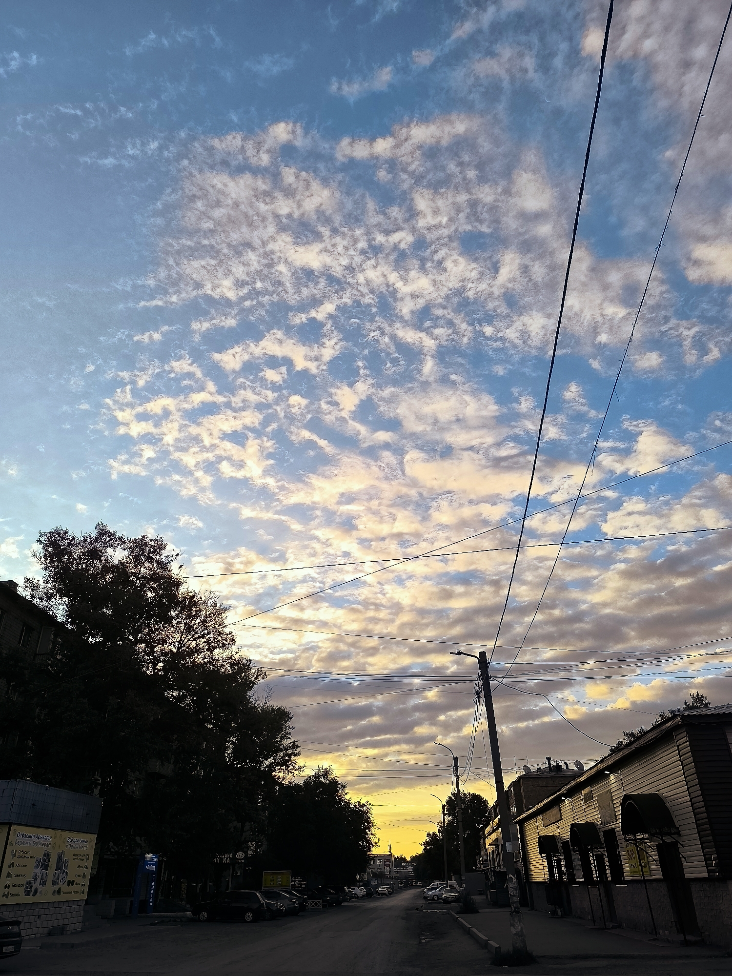 The image depicts a street scene in Qarağandı, Kazakhstan, during what appears to be sunset or sunrise. The sky is filled with scattered clouds tinged with hues of yellow, orange, and blue. Several power lines are visible crisscrossing the sky. On the left side of the street, there are buildings and a large tree silhouetted against the bright sky. On the right side, there is a low-rise building with a distinct facade. Several parked cars are visible along the street, and there are streetlights and a utility pole. The overall impression is a view of an urban environment under a dynamic sky.