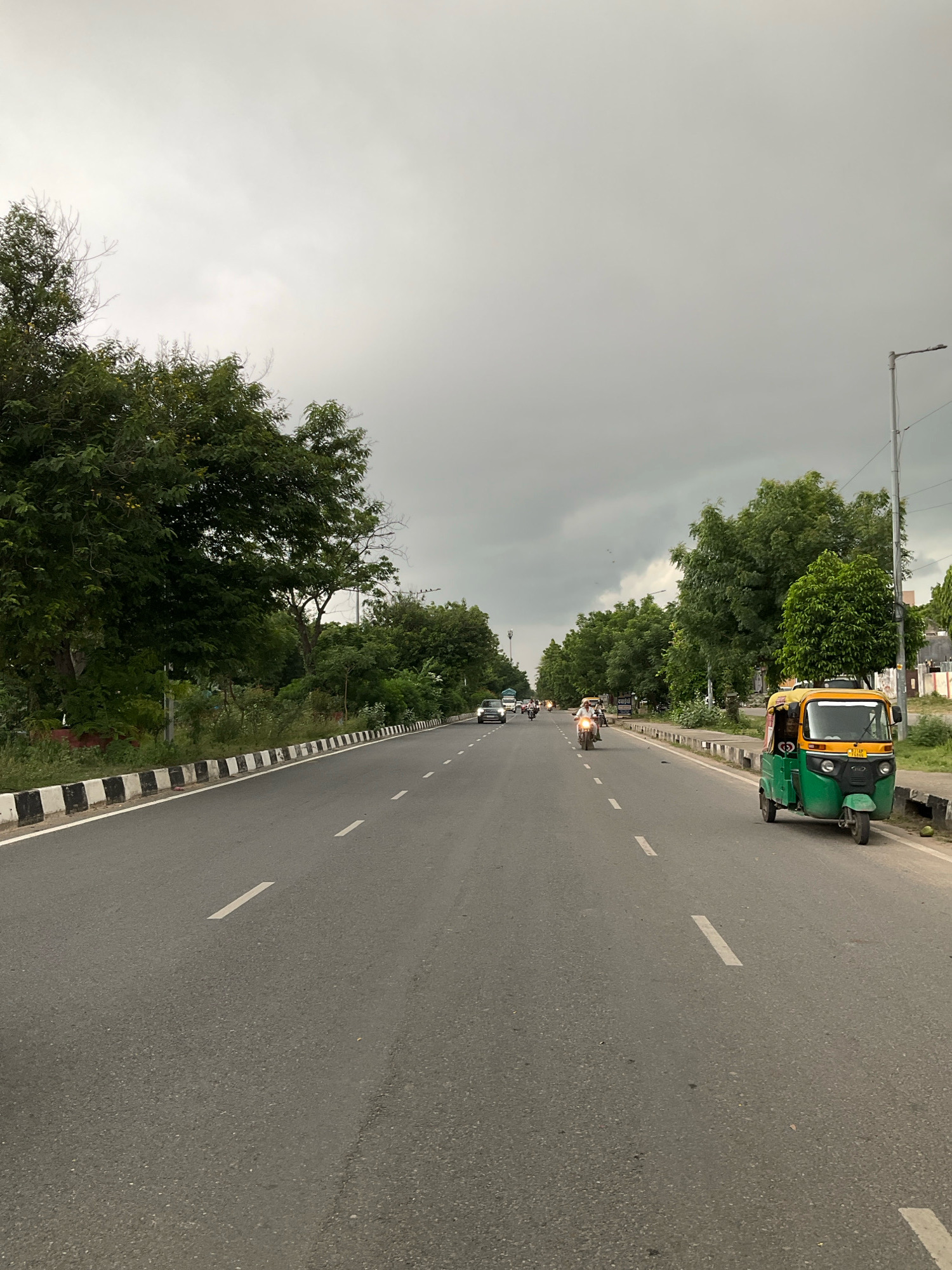 An exterior shot captures a cloudy, overcast day on a multi-lane asphalt road in Jaipur Municipal Corporation, India. The road features white dashed lane markers. On the right side of the road, a green and yellow auto-rickshaw is parked, with a rider visible in the driver's seat. Beyond the auto-rickshaw, a street lamp stands tall. A black and white striped barrier delineates the edge of the road. Lush green trees line both sides of the road. In the distance, several vehicles, including cars and motorcycles, are traveling on the road, with a few individuals on motorcycles. The sky is heavily clouded, obscuring any clear view of the sun. The overall scene depicts a typical road in a moderately busy urban or semi-urban environment.