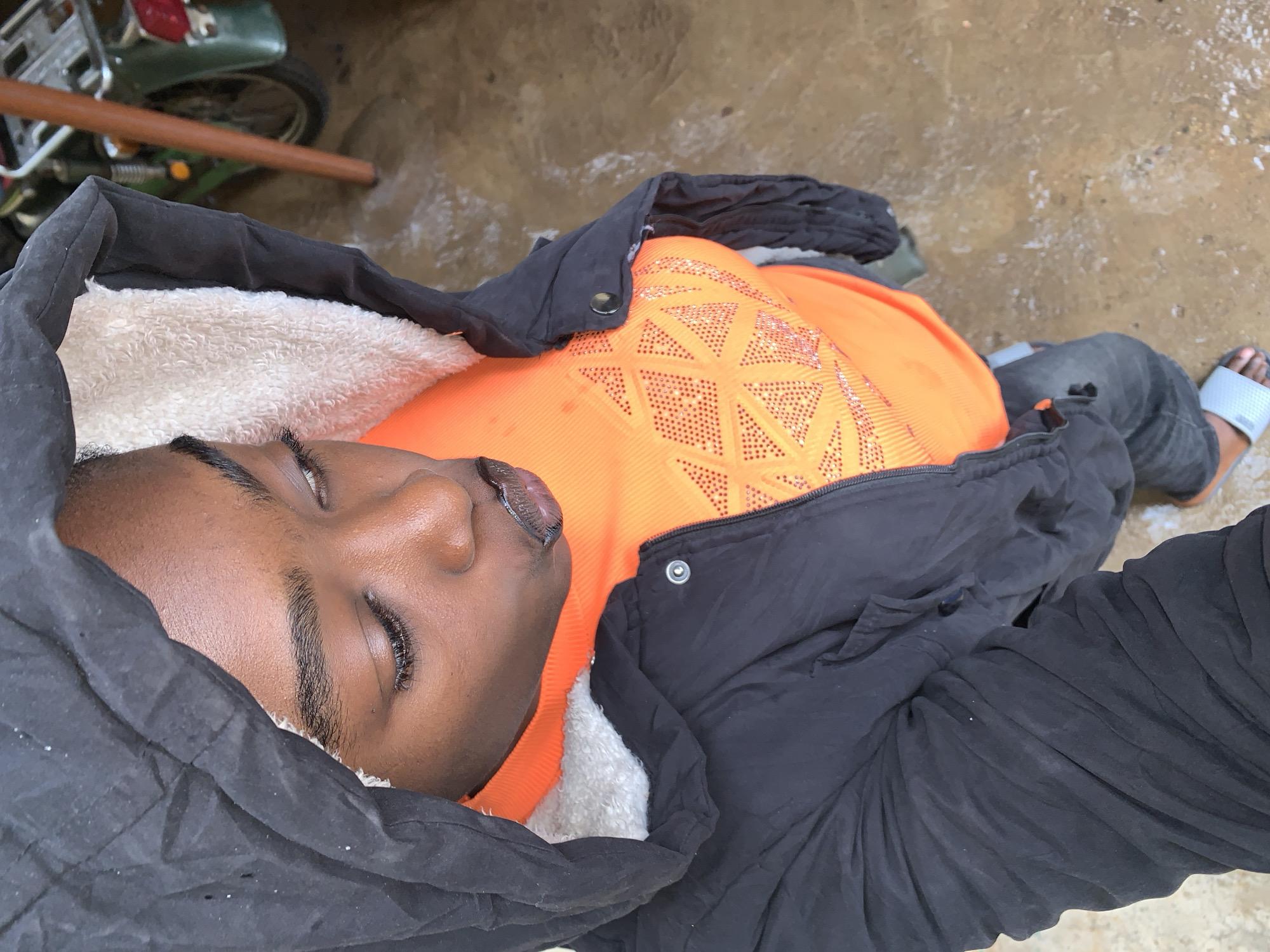 A medium-shot, overhead, indoor photograph depicts a young Black individual with their hood up, looking down and to the right with lips pursed. They are wearing an orange, long-sleeved shirt adorned with a diamond-patterned embellishment in shades of orange and silver. Over this, they wear a black puffer jacket with a white, furry lining visible around the hood and collar. Dark wash jeans with a rip on the left thigh are visible, along with light blue slide sandals. The individual is standing on a wet, concrete surface that appears to be outdoors, possibly a driveway or covered patio, with a green motorcycle and a brown pole visible in the background. The scene suggests a cool or wet environment.