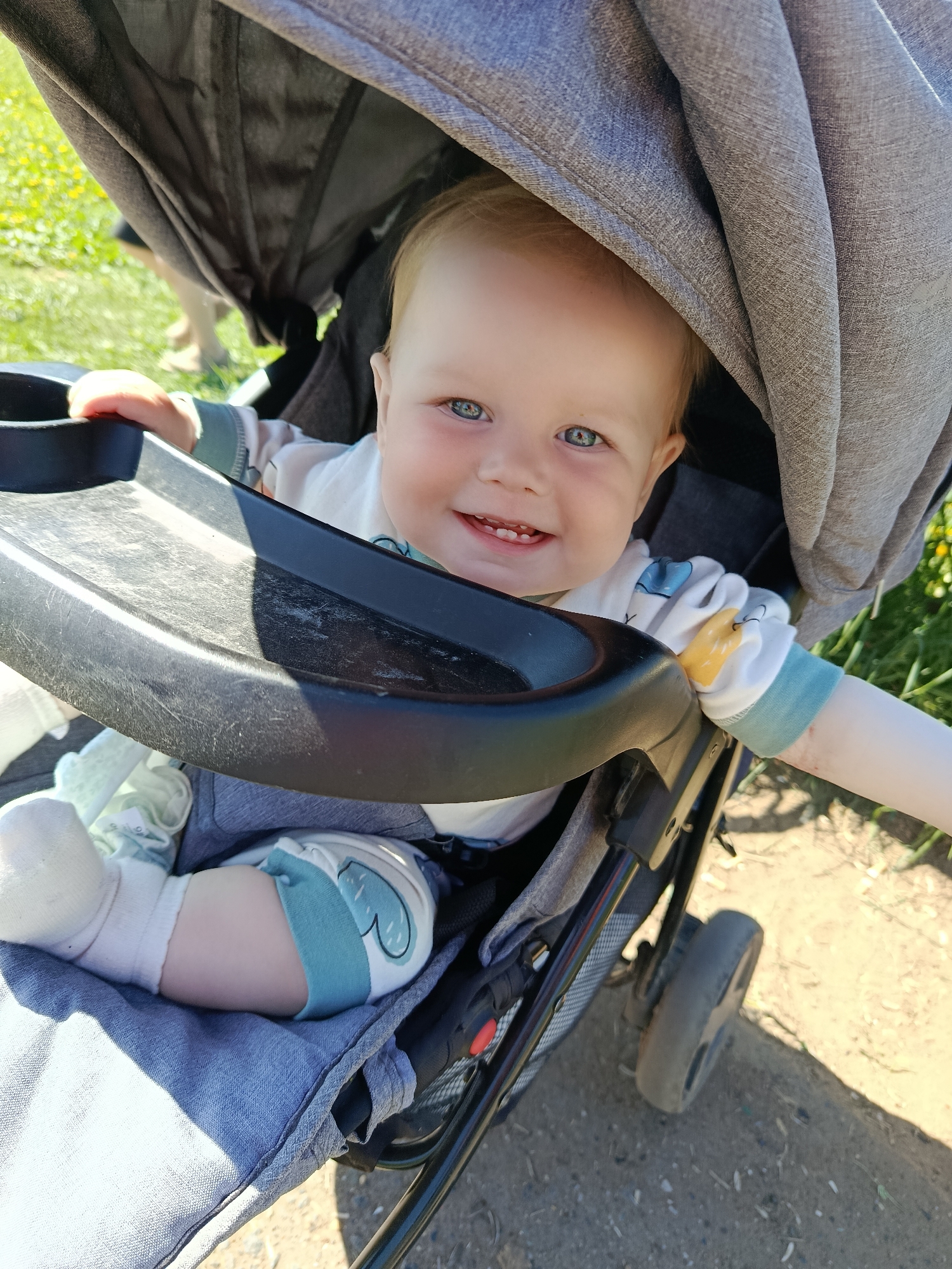A baby is sitting in a stroller. The baby is fair-skinned with light blonde hair and blue eyes, and is smiling, showing a few teeth. The baby is wearing a white outfit with light blue and yellow accents. The stroller is gray and black and has a tray in front of the baby. The stroller's sunshade is partially extended. The stroller is on a dirt path in a grassy area.
It's impossible to determine the location as the city of Фоки, Russia, based on visual information.