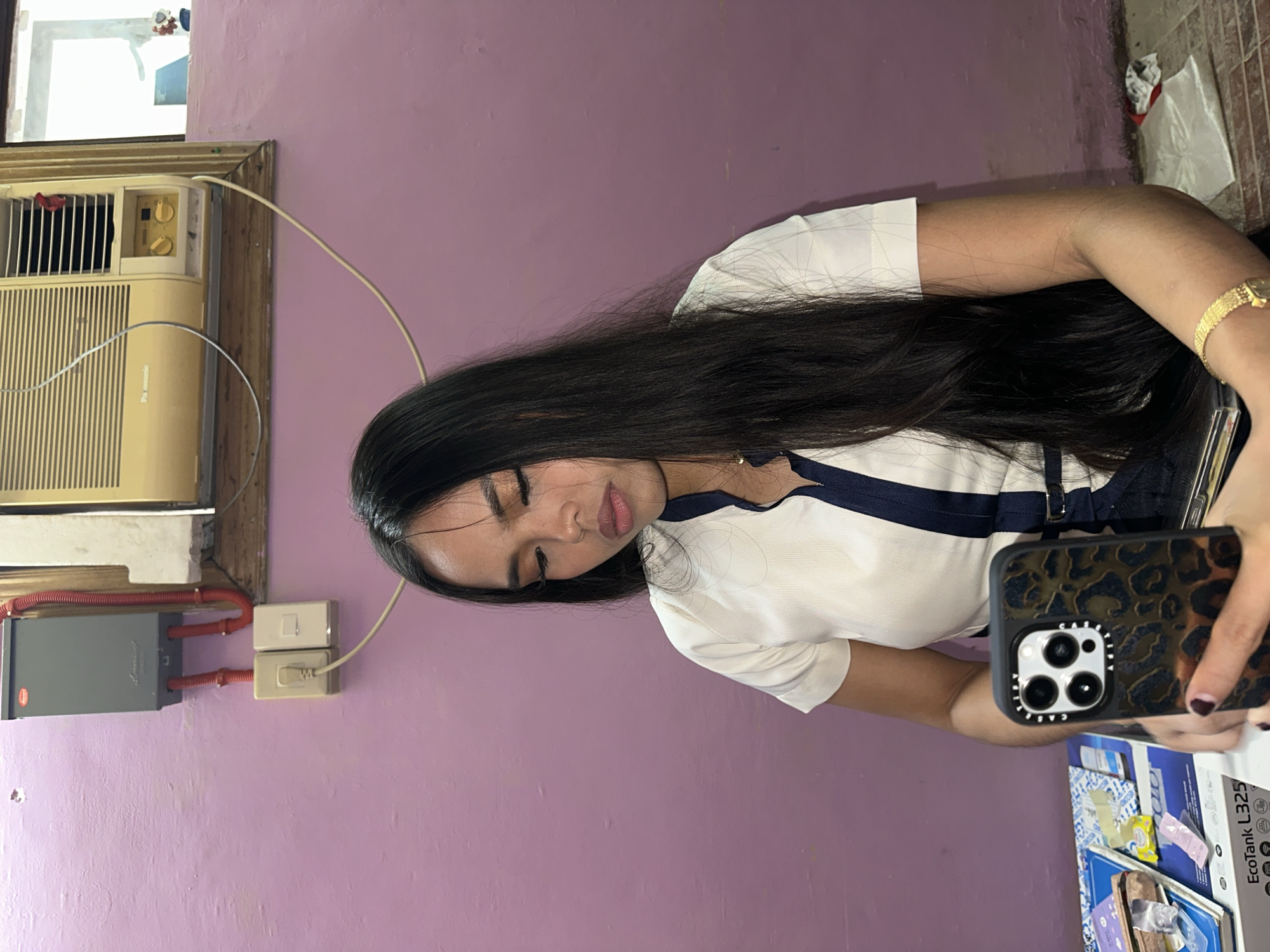 A young woman with long, dark hair is sitting in a room with a purple wall. She is looking down at her phone, which has a leopard-print case. She is wearing a white shirt with a dark blue collar and trim. In the background, there is an air conditioner mounted high on the wall, and an electrical panel with red conduits. To her left, there are stacks of books and papers on a surface, and a box that reads "EcoTank L325". The floor appears to be tiled. The scene suggests an indoor setting in a residential or office environment in Davao City, Philippines.