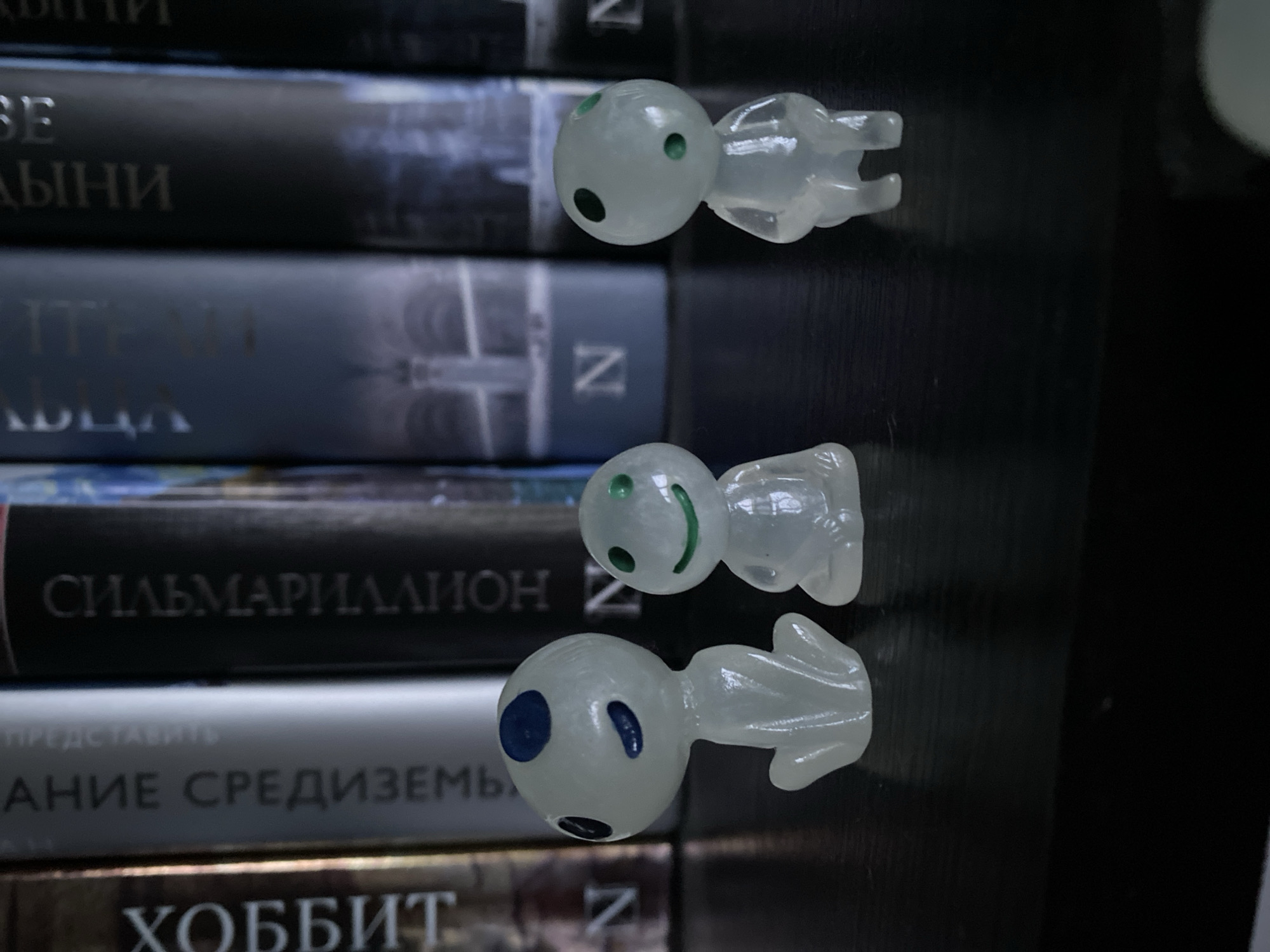 The image shows a stack of books with three small, translucent, figurine-like objects placed on top. The books' spines are visible, with titles in what appears to be the Cyrillic alphabet. One book clearly reads "Сильмариллион" (Silmarillion) and another "Хоббит" (Hobbit). The figurines are vaguely humanoid in shape, featuring round "heads" with dark, circular markings resembling eyes and mouths. The environment appears to be indoors, potentially a bookshelf or similar display. There is nothing in the image to indicate it was taken in Novokuznetsk, Russia.
