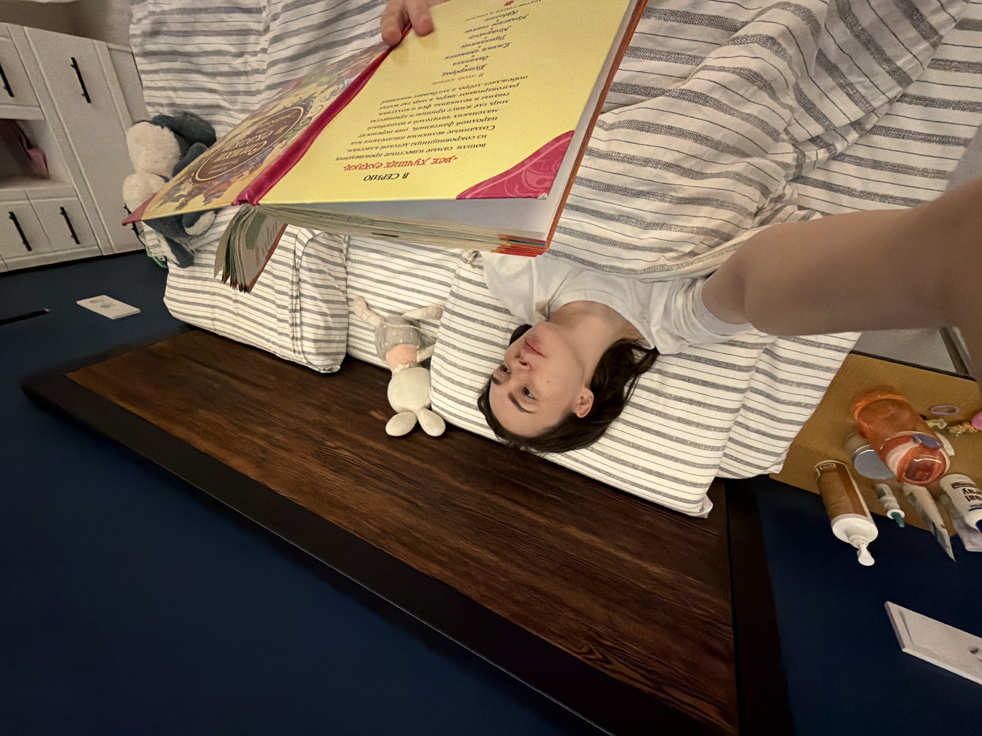A person is lying on a striped bed, holding a large, open book with Russian text visible. The book's spine is red, and the pages are yellow. To the person's left, there is a small stuffed animal and a wooden bedside table with a dark border. Various personal care items, like tubes and bottles, are on a blue surface next to the bed. In the background, there is a white cabinet or shelving unit. The picture is taken from an unusual angle, making the scene appear tilted. The location could be a home in the city of Chaikovsky, Russia, given the Russian text in the book.