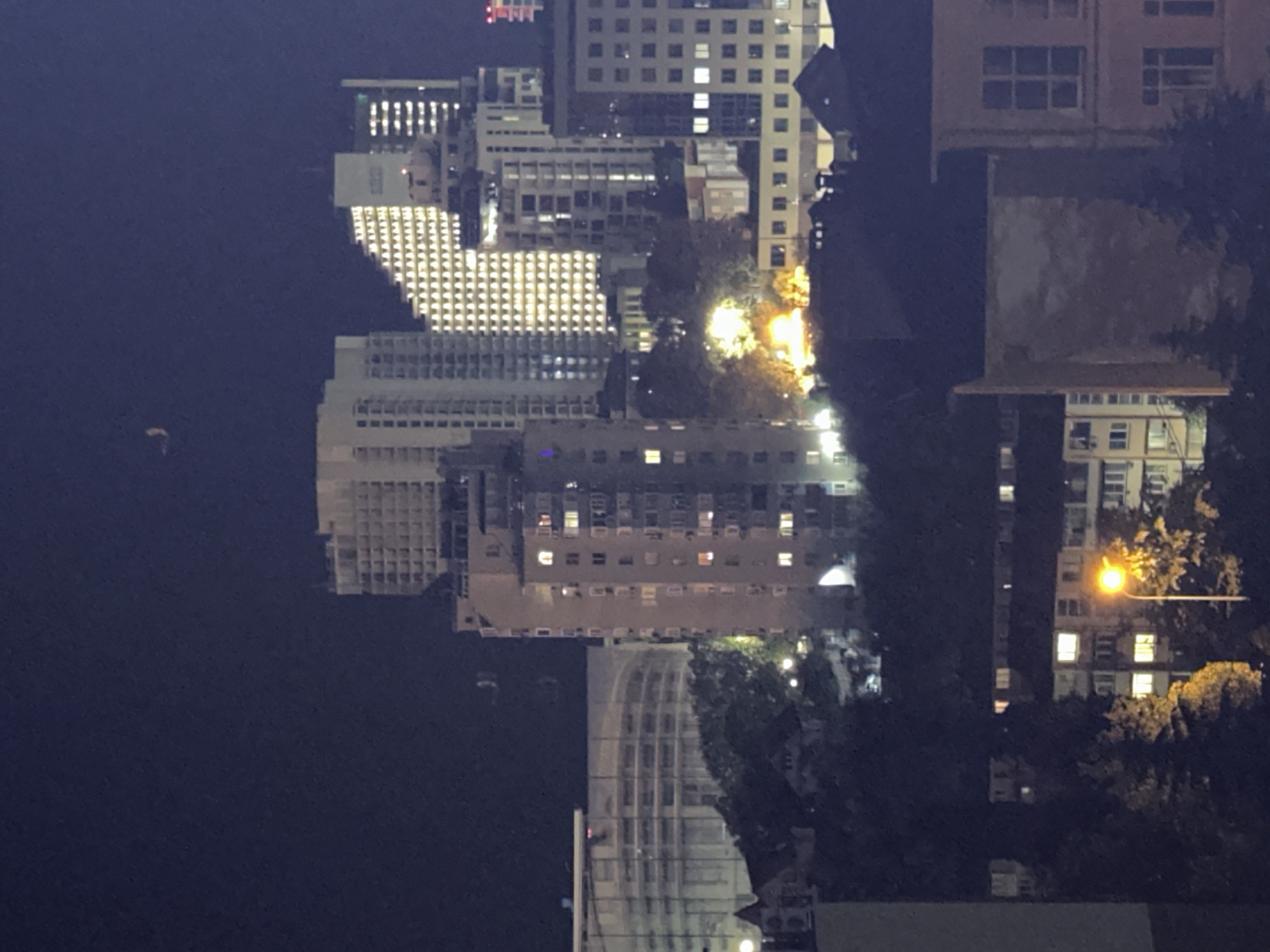 The image depicts a nighttime cityscape with a cluster of modern buildings of varying heights and architectural styles, illuminated by interior lights and streetlights. A crescent moon is visible in the dark sky. The buildings are a mix of residential and possibly commercial structures, some featuring balconies and many windows. The foreground shows a streetlamp casting a warm glow on trees and the lower levels of some buildings, suggesting a residential area within the city. The overall impression is of an urban environment at night, likely from an elevated vantage point. The context provided states the location is Sochi, Russia.