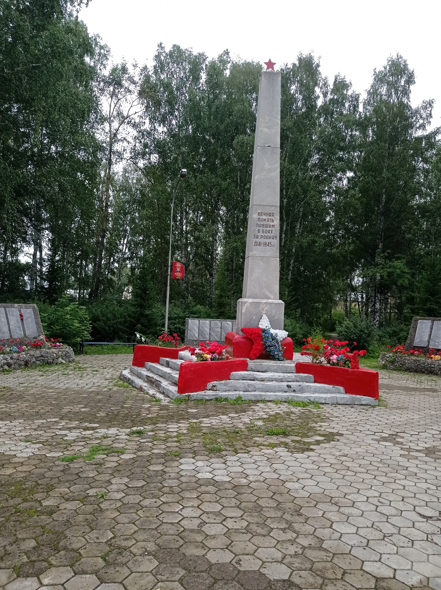 The image depicts a war memorial in the city of Degtyarsk, Russia. The central feature is a tall, grey obelisk topped with a red star. Inscriptions in Russian are visible on the obelisk, reading "ETERNAL MEMORY TO THOSE WHO FELL IN BATTLE FOR THE HOMELAND 1941-1945". At the base of the obelisk, a red, stepped platform supports a wreath of red and blue flowers.
To the left of the obelisk, another war memorial is visible, constructed from stones and concrete, with names inscribed on plaques. This memorial is also adorned with flowers. To the right, a similar stone memorial with plaques is partially visible, also decorated with red flowers.
The memorial area is paved with hexagonal grey interlocking concrete stones, with some grass growing between them. A flagpole stands to the right of the obelisk, bearing a red banner with a symbol. The background is dominated by a dense stand of tall green trees, suggesting a park or wooded area. The sky is overcast. No people are present in the scene. The overall impression is one of solemn remembrance.