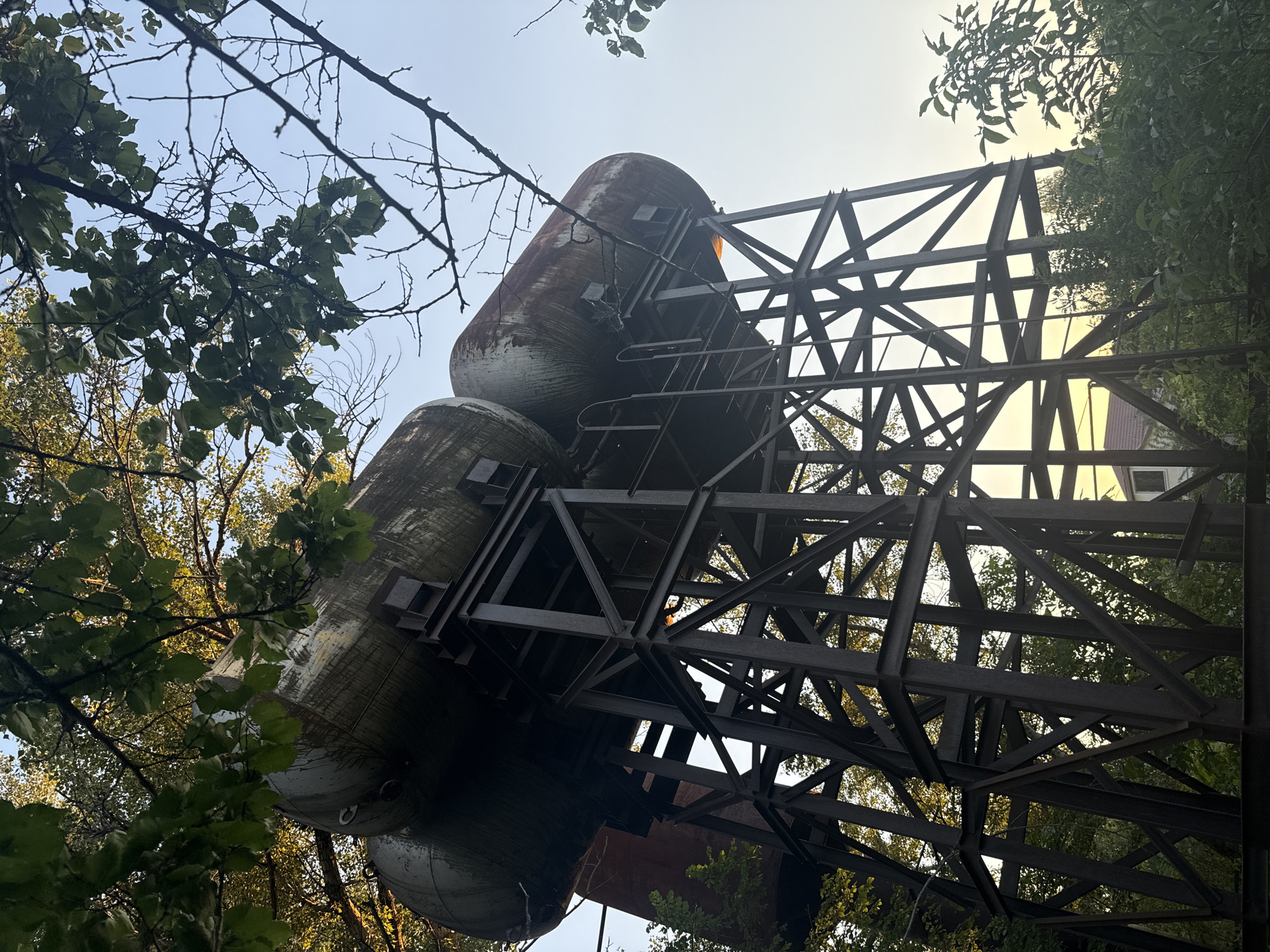 This is a low-angle, medium shot of an abandoned industrial water tower structure with three large, cylindrical tanks on top. The tanks are weathered, with visible rust and streaks of discoloration. They are supported by a tall, dark, metal framework constructed of I-beams and diagonal bracing. The structure is partially obscured by lush green foliage and tree branches, with dappled sunlight filtering through the leaves. In the background, the sky is clear and pale blue, with a hint of sunset or sunrise glow. A portion of a residential building with a brown roof is visible through the trees and the metal structure. The scene is set in a naturalistic environment within the city of Obukhivka, Ukraine. There are no people or animals present in the image.