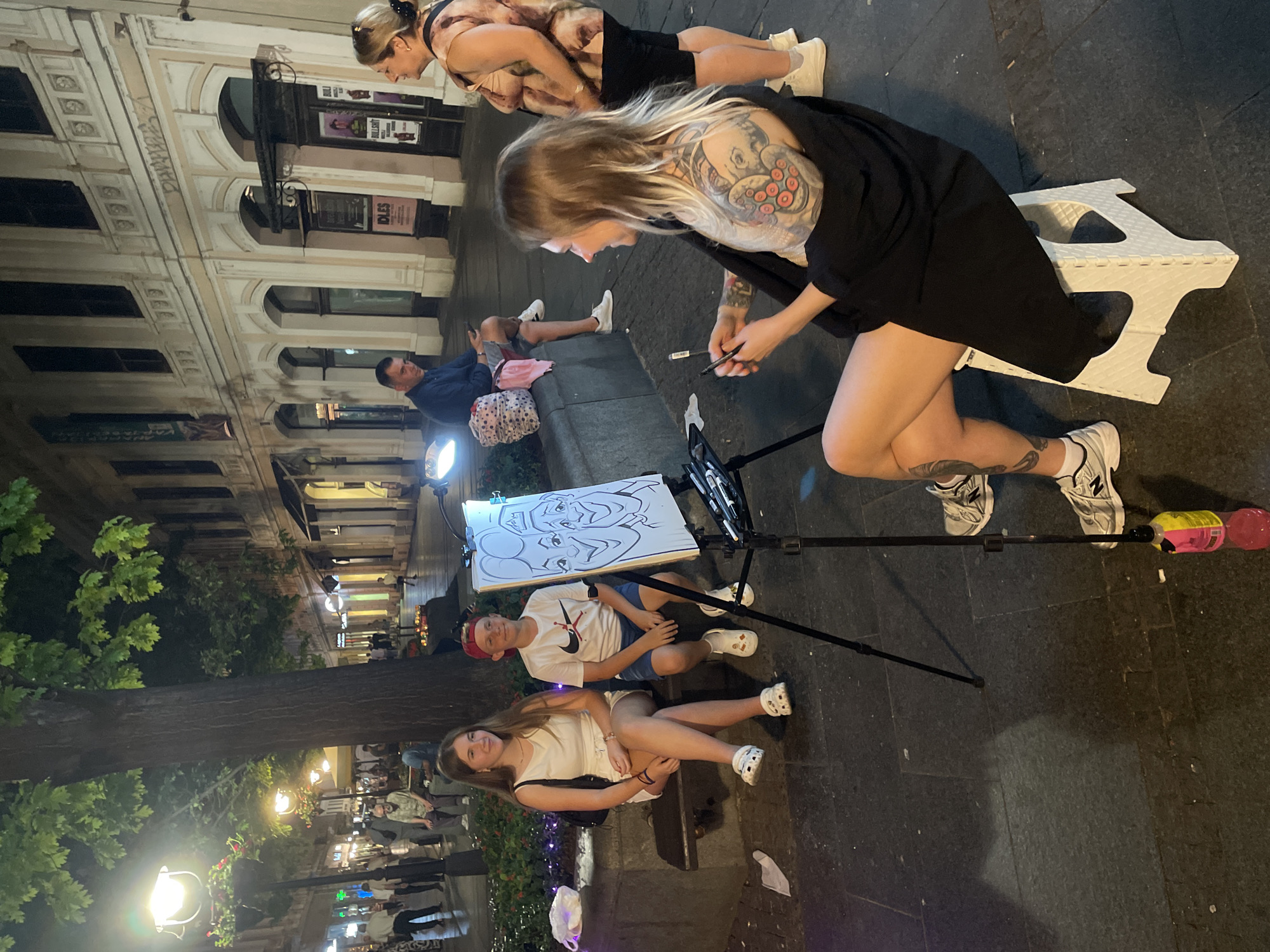 This image depicts a street scene at night, likely in Belgrade, Serbia, characterized by buildings with arched windows and illuminated storefronts lining a cobblestone street. In the foreground, a woman with extensive tattoos on her arm is seated on a white stool, facing away from the camera and towards a caricature artist's easel. She is holding drawing utensils and appears to be in the process of creating a caricature. The easel displays a partially completed drawing of two faces. To the left of the easel, a young girl and boy are seated on a stone bench. The girl, wearing a white dress and white clogs, is looking towards the artist. The boy, wearing a white t-shirt and blue shorts, is also looking towards the artist. In the background, other people are walking along the street, and a man is seated on a bench further down. Streetlights and trees add to the ambient lighting of the scene. A pink and yellow plastic bottle is attached to the tripod supporting the easel.